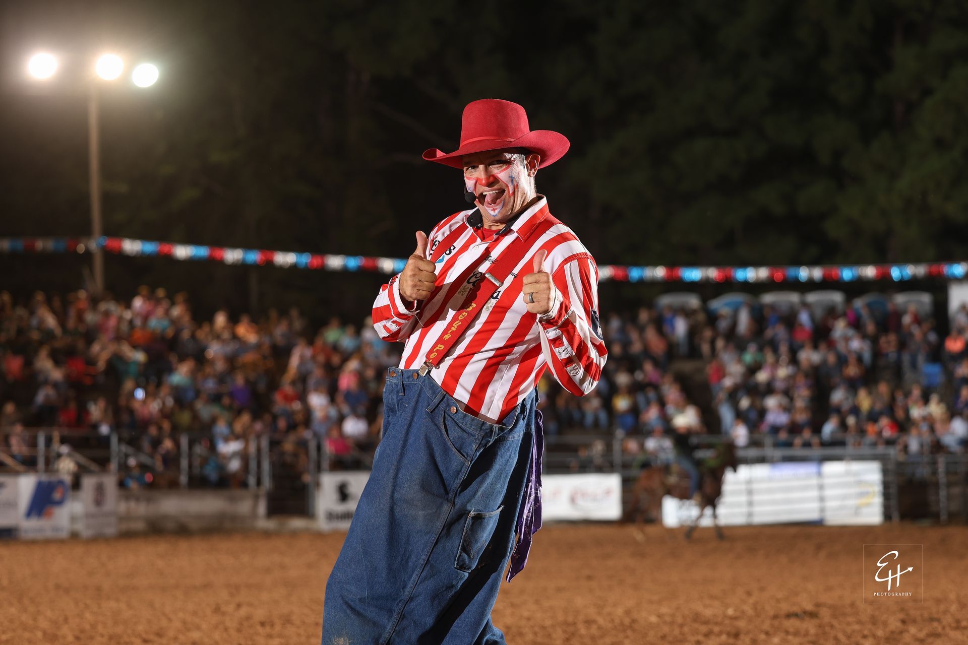 Clown in red hat and striped shirt performing in a rodeo arena at night, with a cheering crowd behind him.