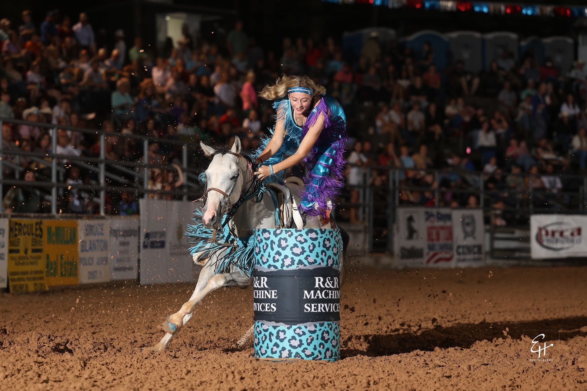 Rider on a white horse racing around a blue barrel in a rodeo arena, crowd in the background