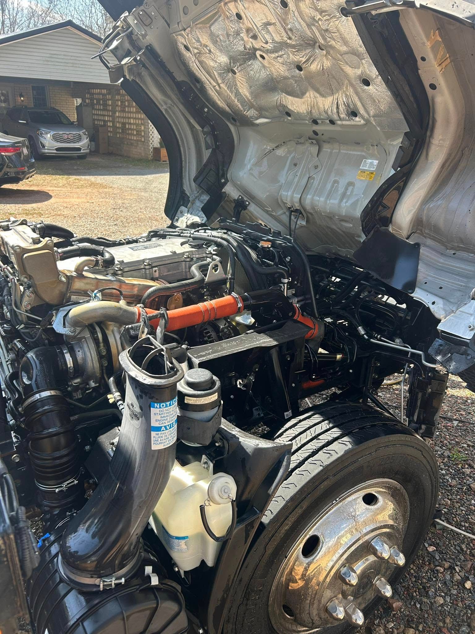 Open truck engine bay with hood raised, showing black hoses and metal components, front wheel visible.