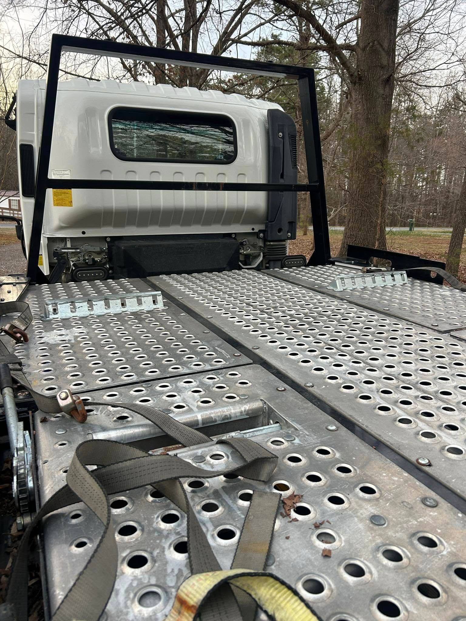 Empty flatbed tow truck with silver metal bed and white cab parked outdoors under trees