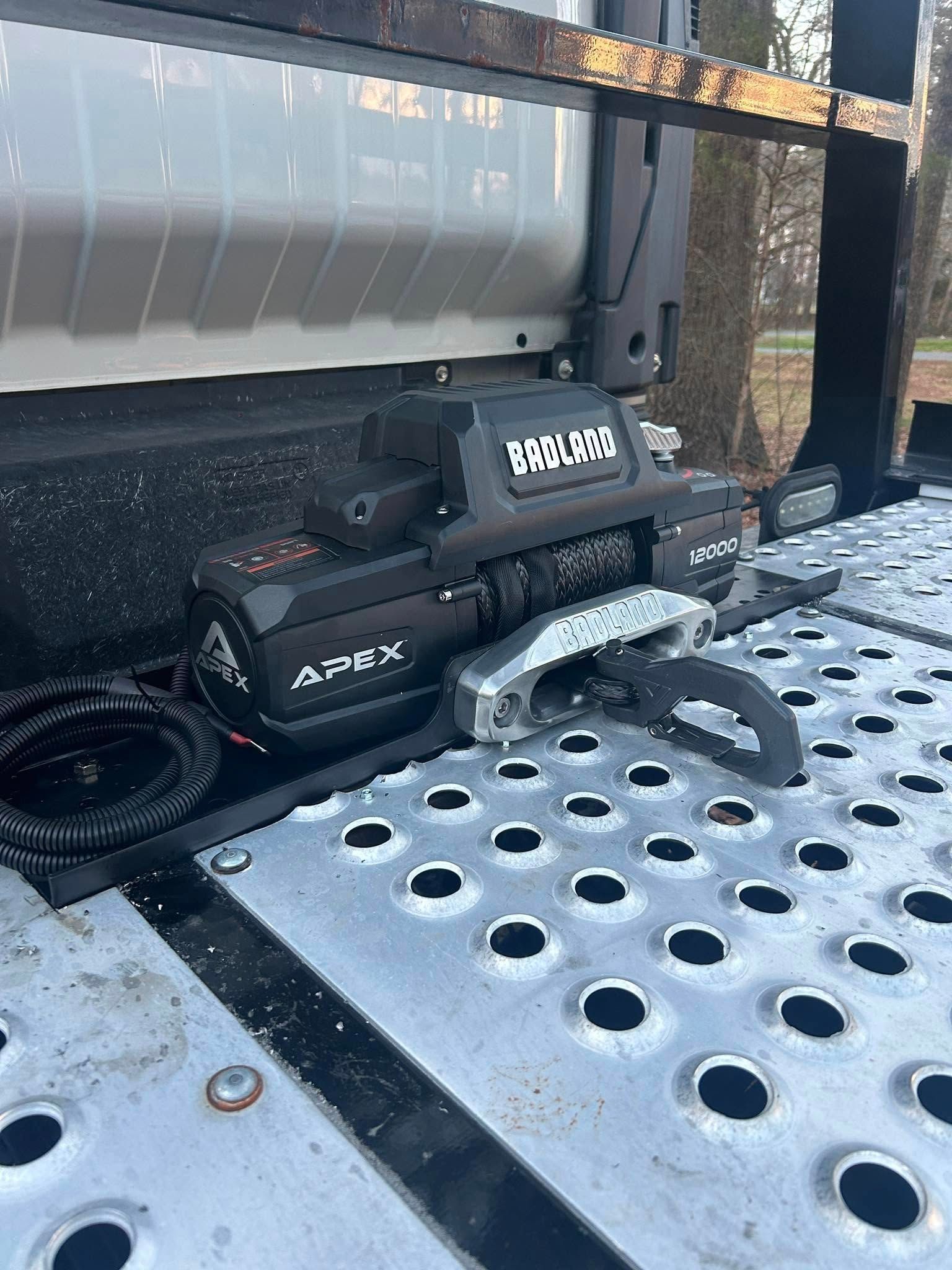 A black ATV winch mounted on a perforated metal trailer deck, with a coiled cable beside it.
