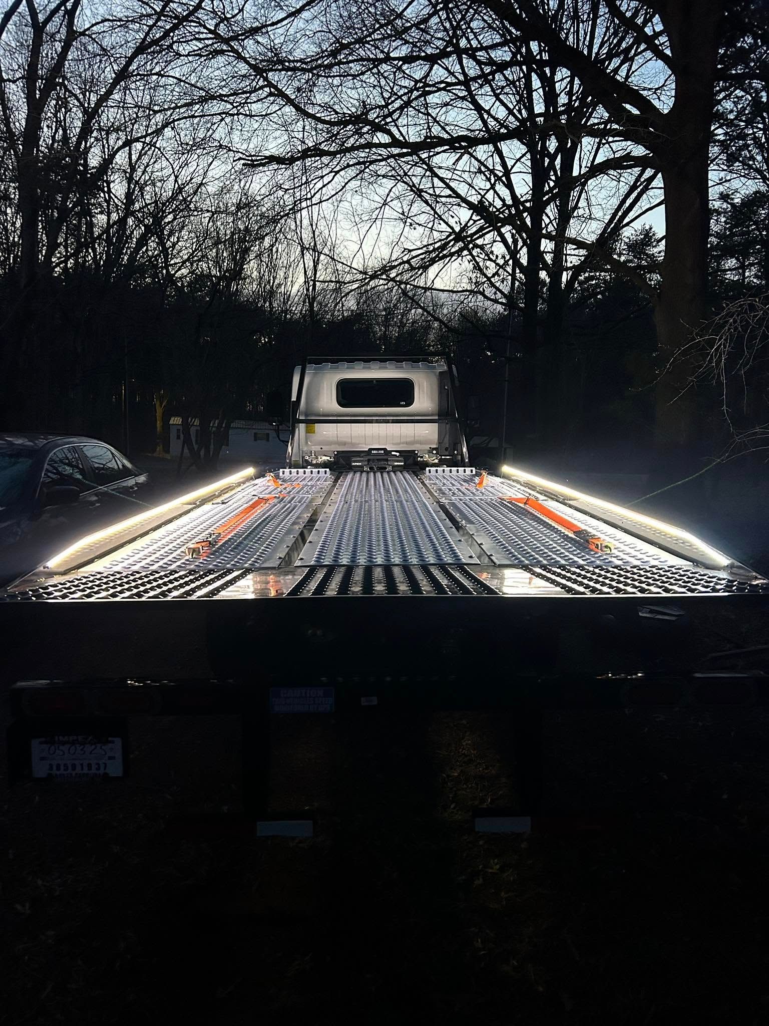 Flatbed tow truck with bright orange side lights parked on a road at dusk, trees silhouetted behind.