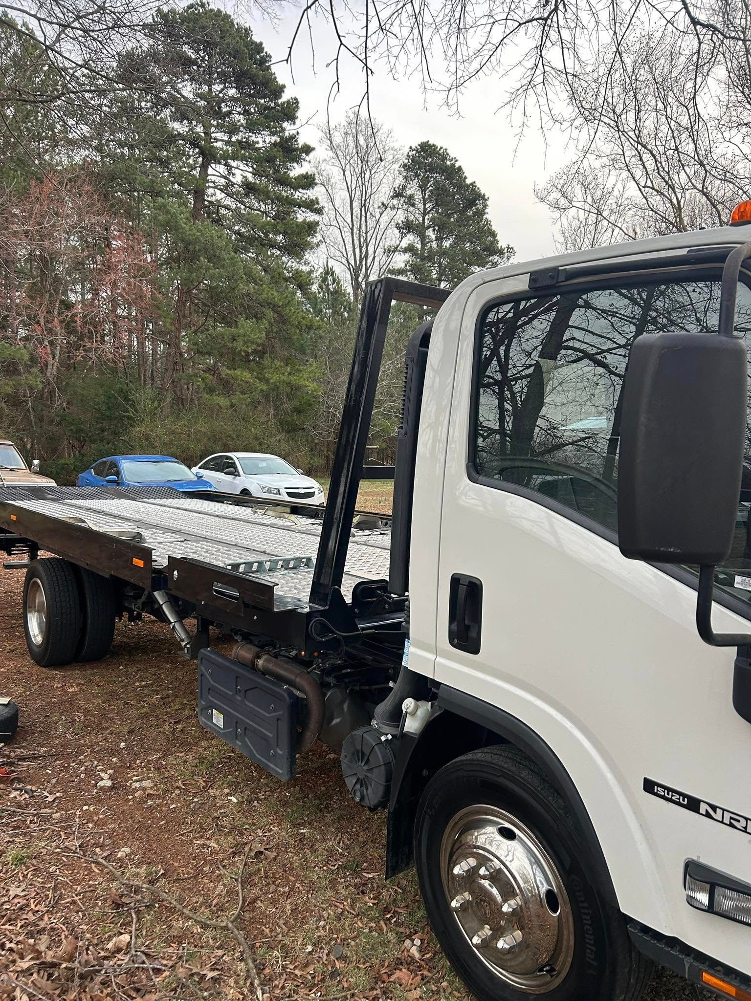 White flatbed tow truck parked on a leaf-covered road with trees in the background.