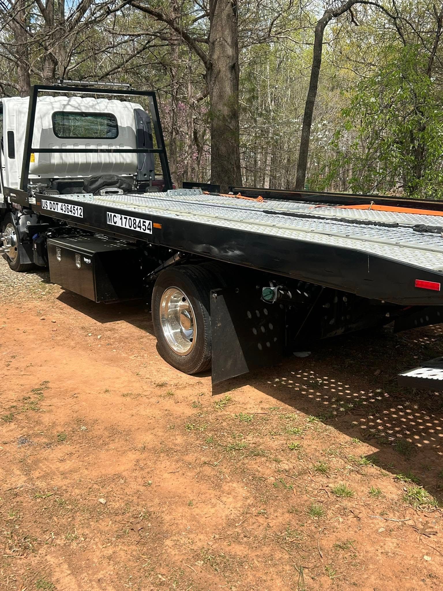 Flatbed tow truck parked on a dirt road beside trees, viewed from the rear side.