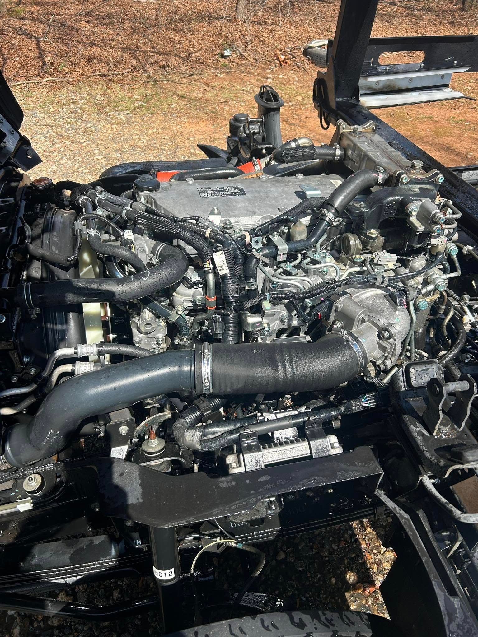 Damaged engine bay with exposed black hoses and components, viewed outdoors on gravel.