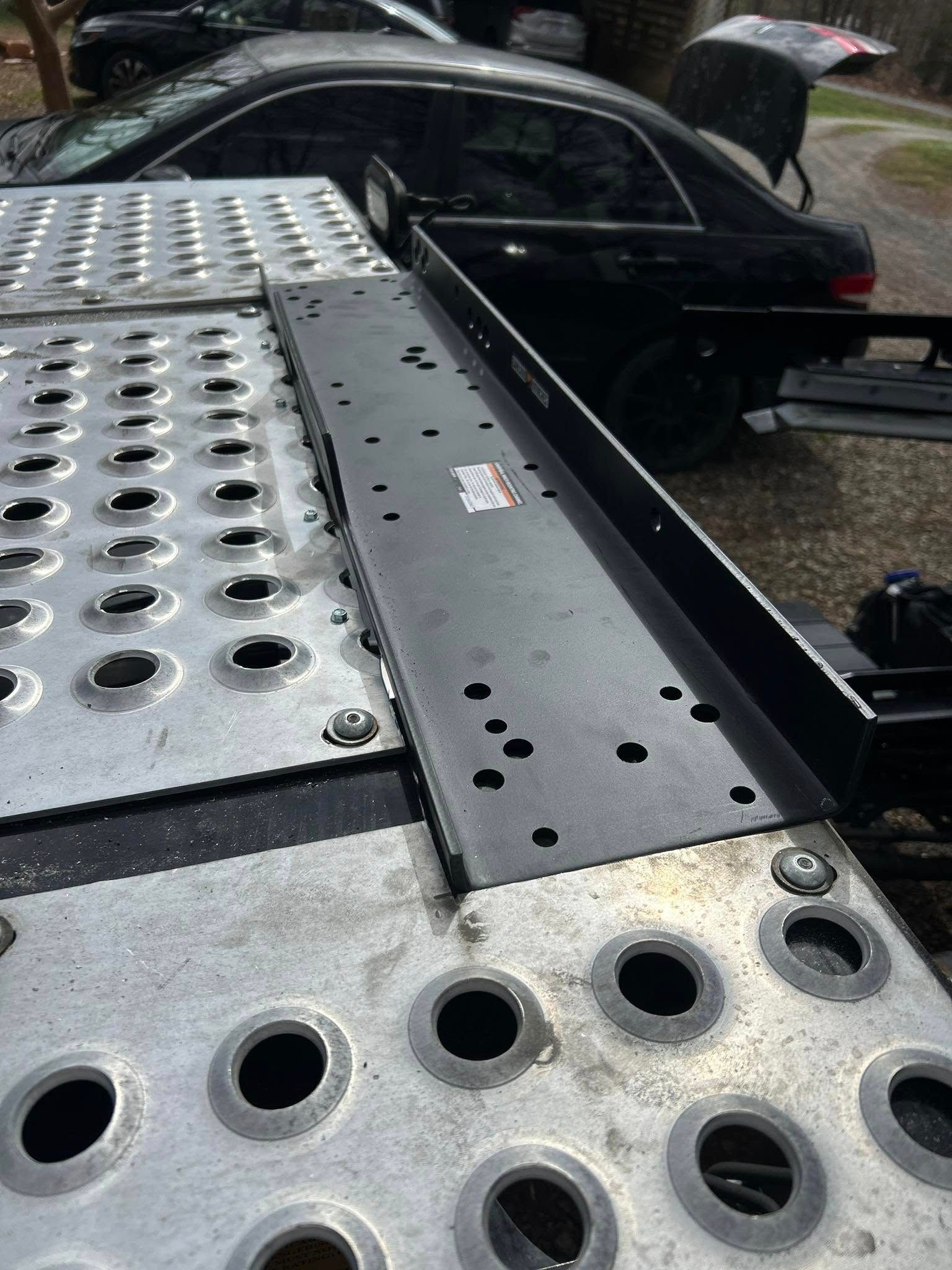 Close-up of a car hauler trailer deck with metal plates and round cutouts beside a black car