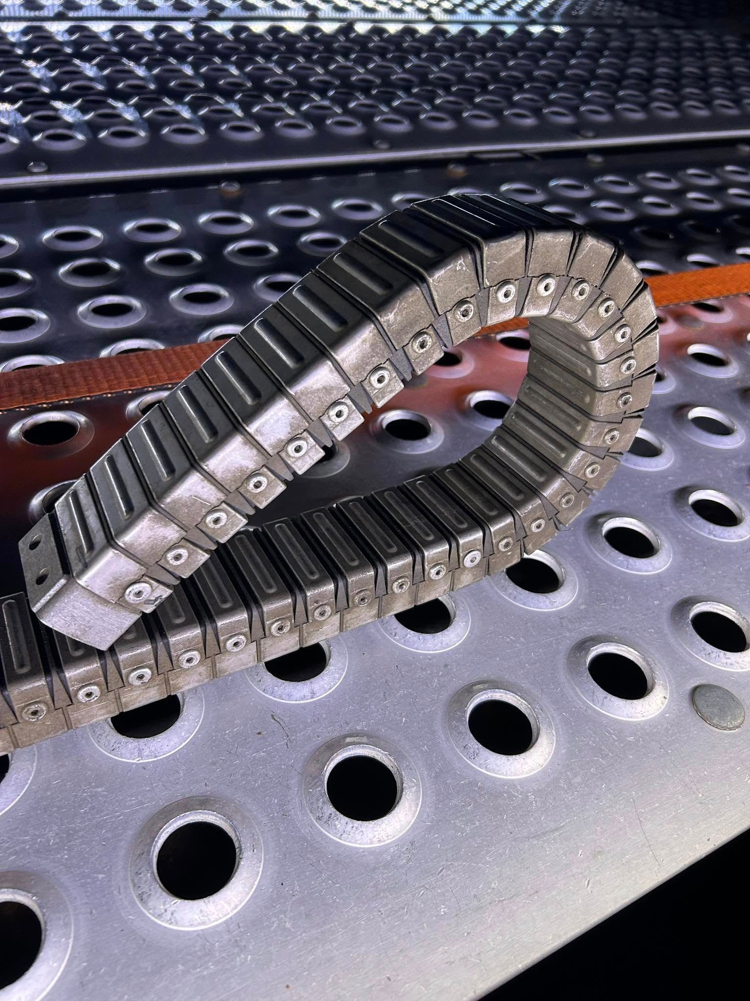 Flexible metal cable carrier loop on a perforated steel surface.