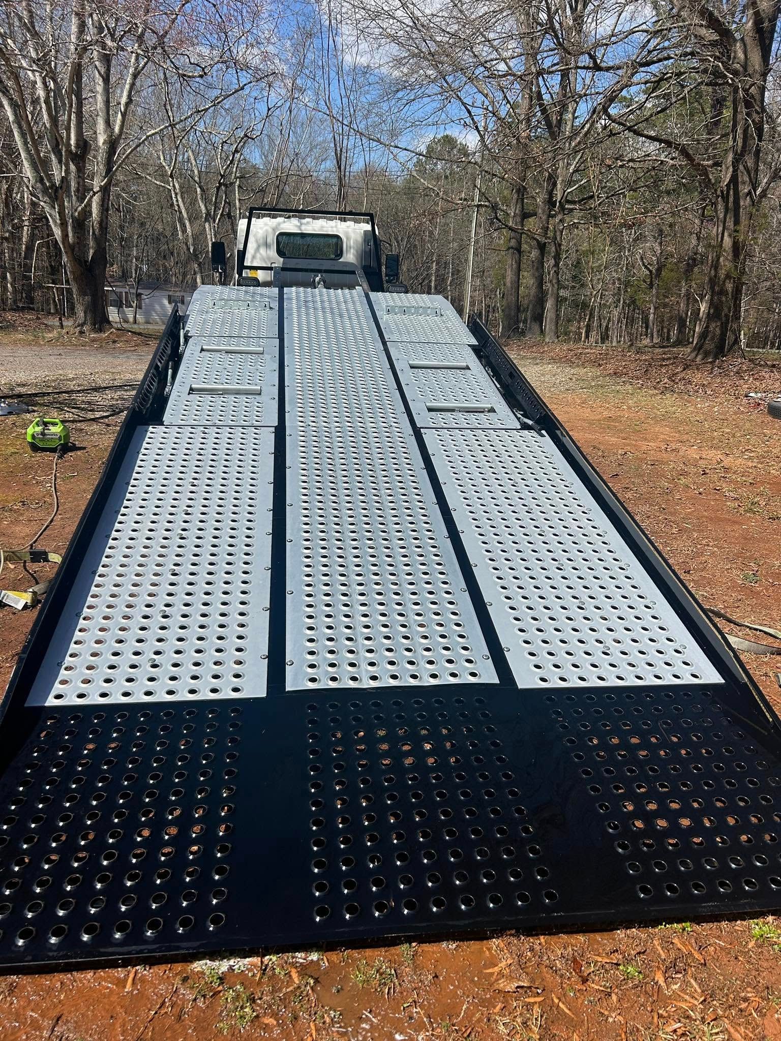 Flatbed trailer ramp on a gravel path in a wooded area, with a truck cab at the top.