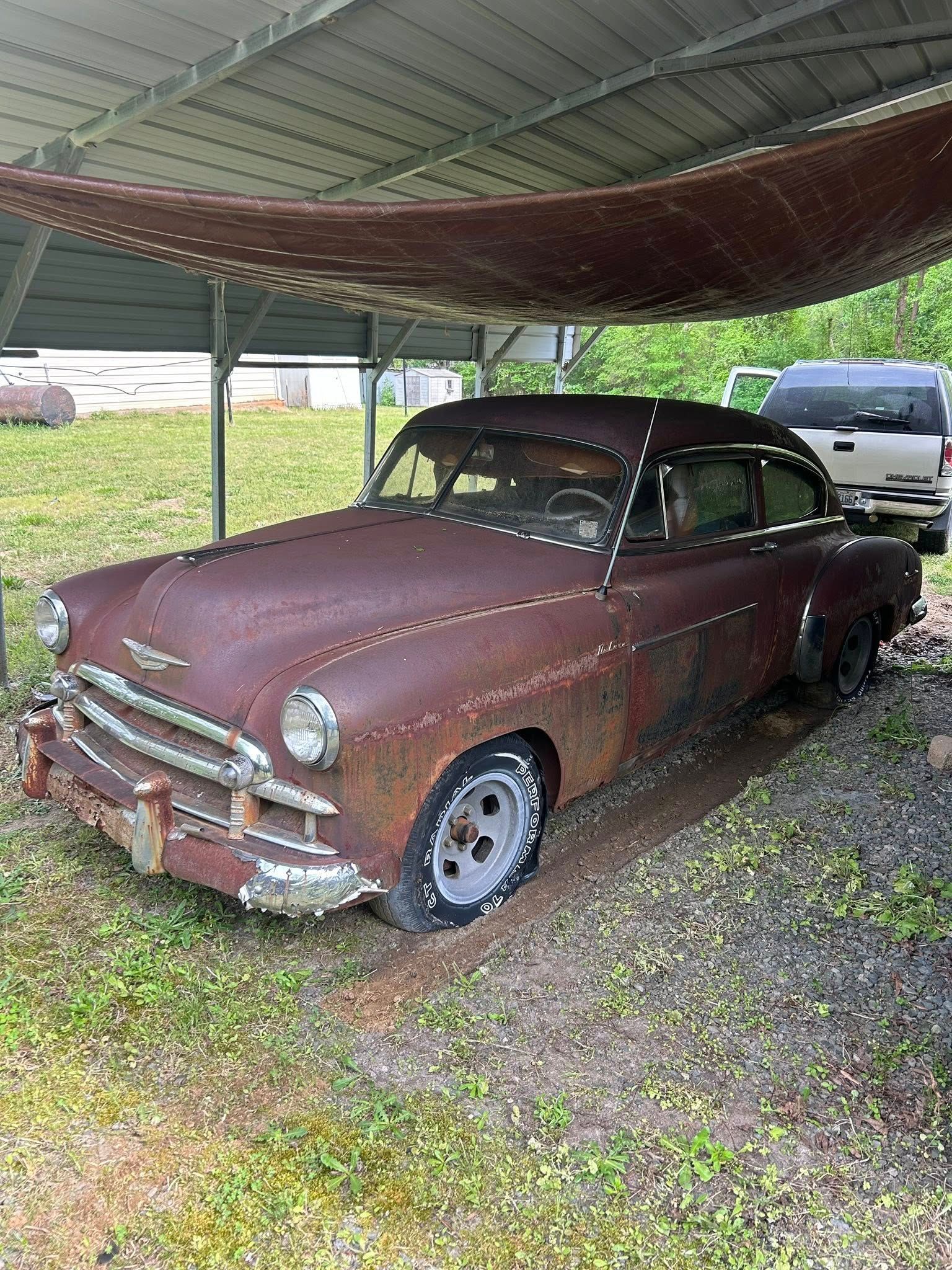 Rusty vintage car parked under a carport on gravel, with a modern vehicle behind it.