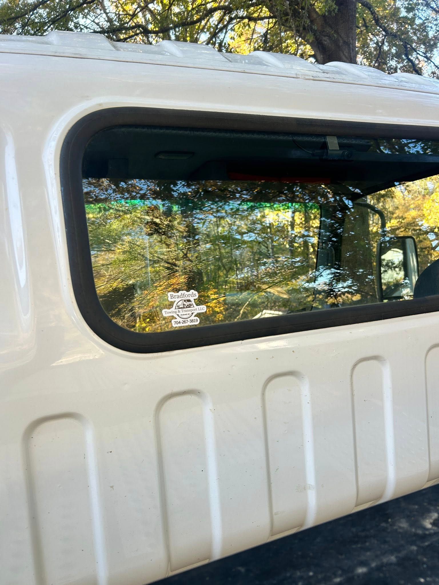 White vehicle side with open rear window reflecting trees and sky outdoors