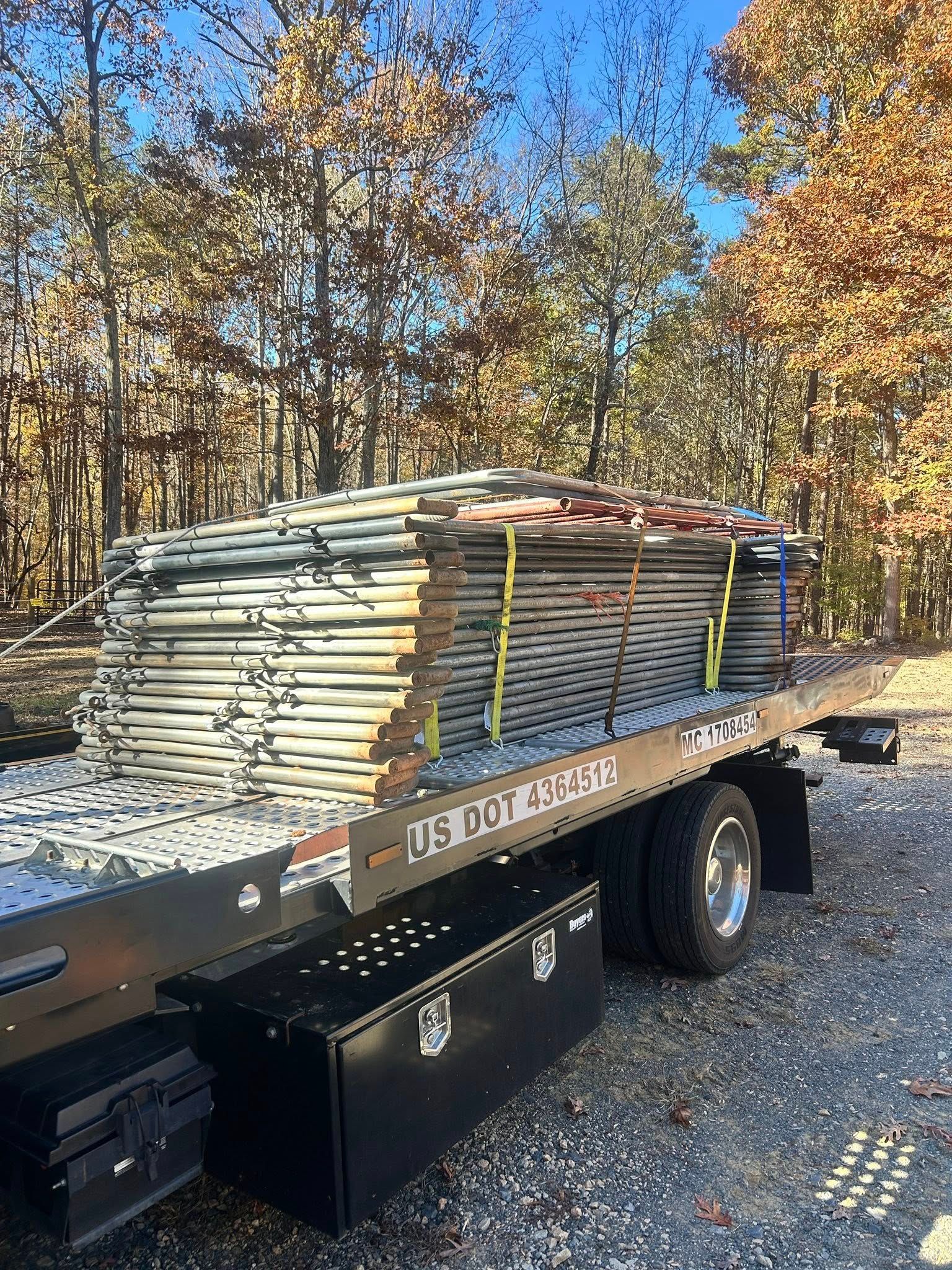 Flatbed trailer loaded with stacked metal pipes, parked on a gravel lot with trees in fall colors.