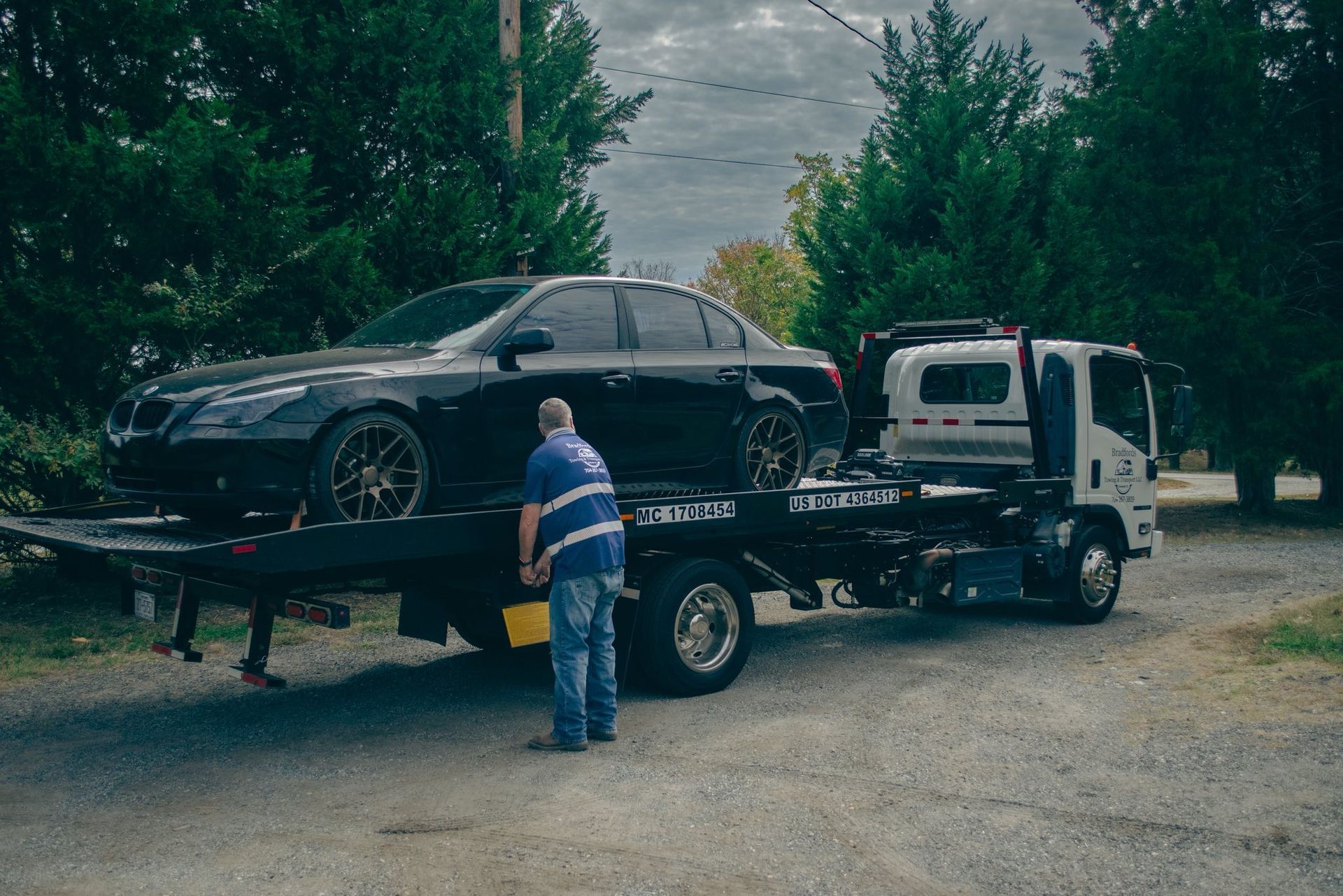 Tow truck loading a black sedan; worker in a striped shirt stands beside it on a gravel road