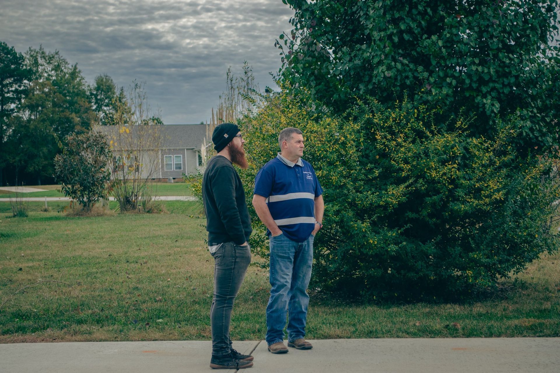 Two people standing on a road beside a large tree in a grassy yard under cloudy skies