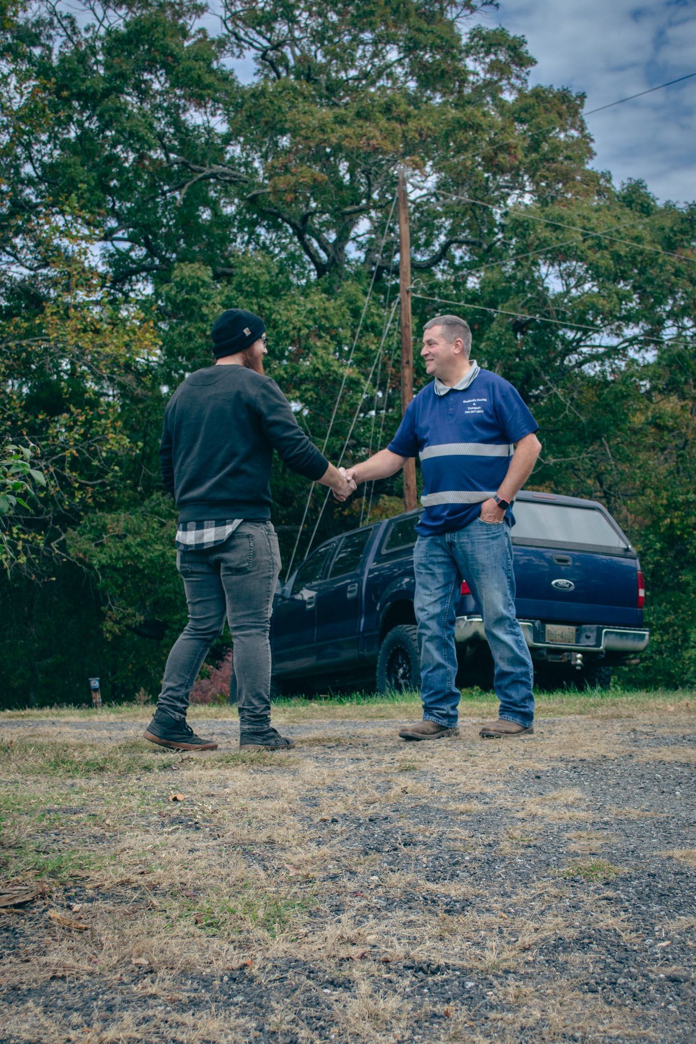 Two people shaking hands on a gravel driveway beside a parked car and trees.