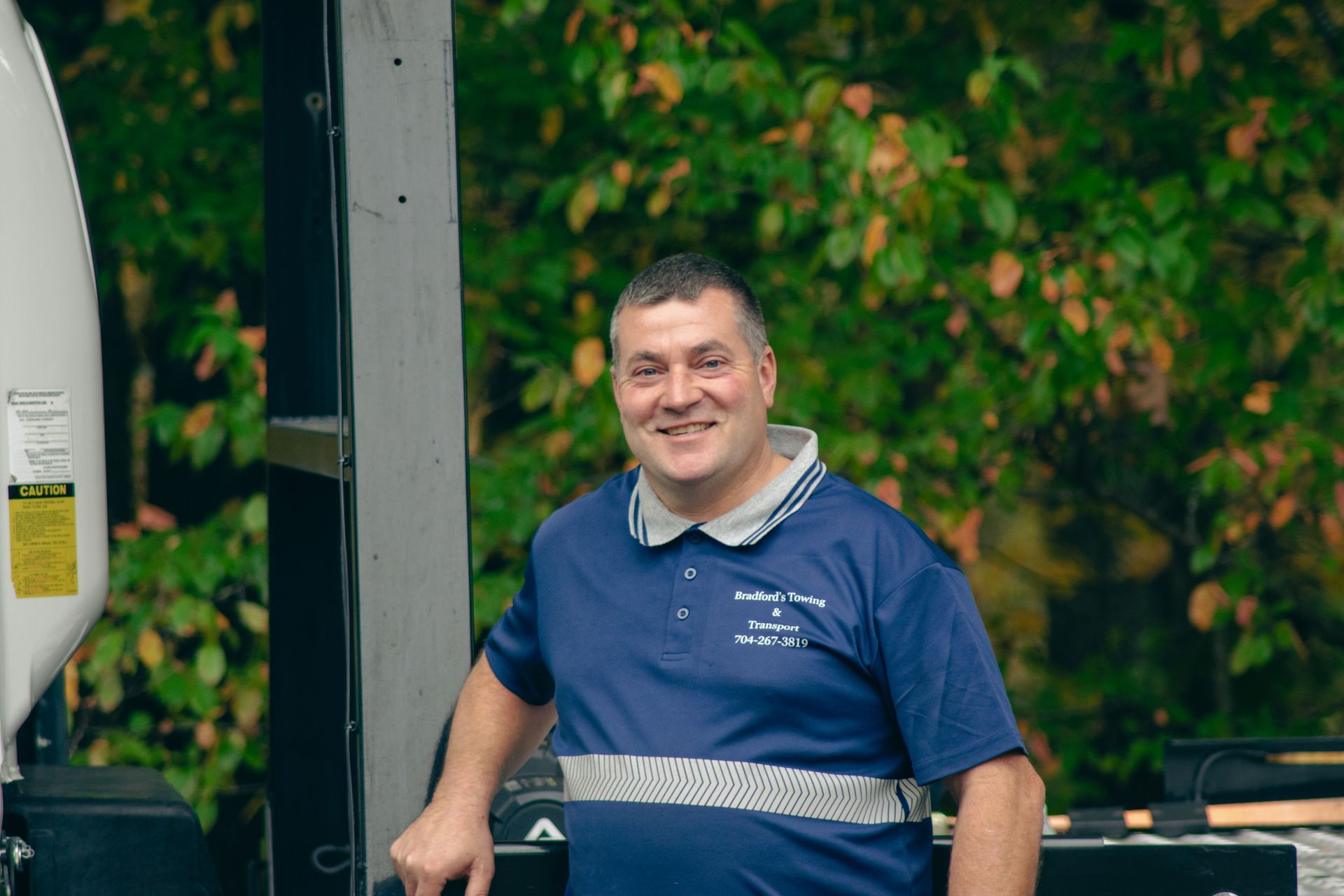 Smiling man in a blue shirt standing by an outdoor grill with leafy greenery behind him