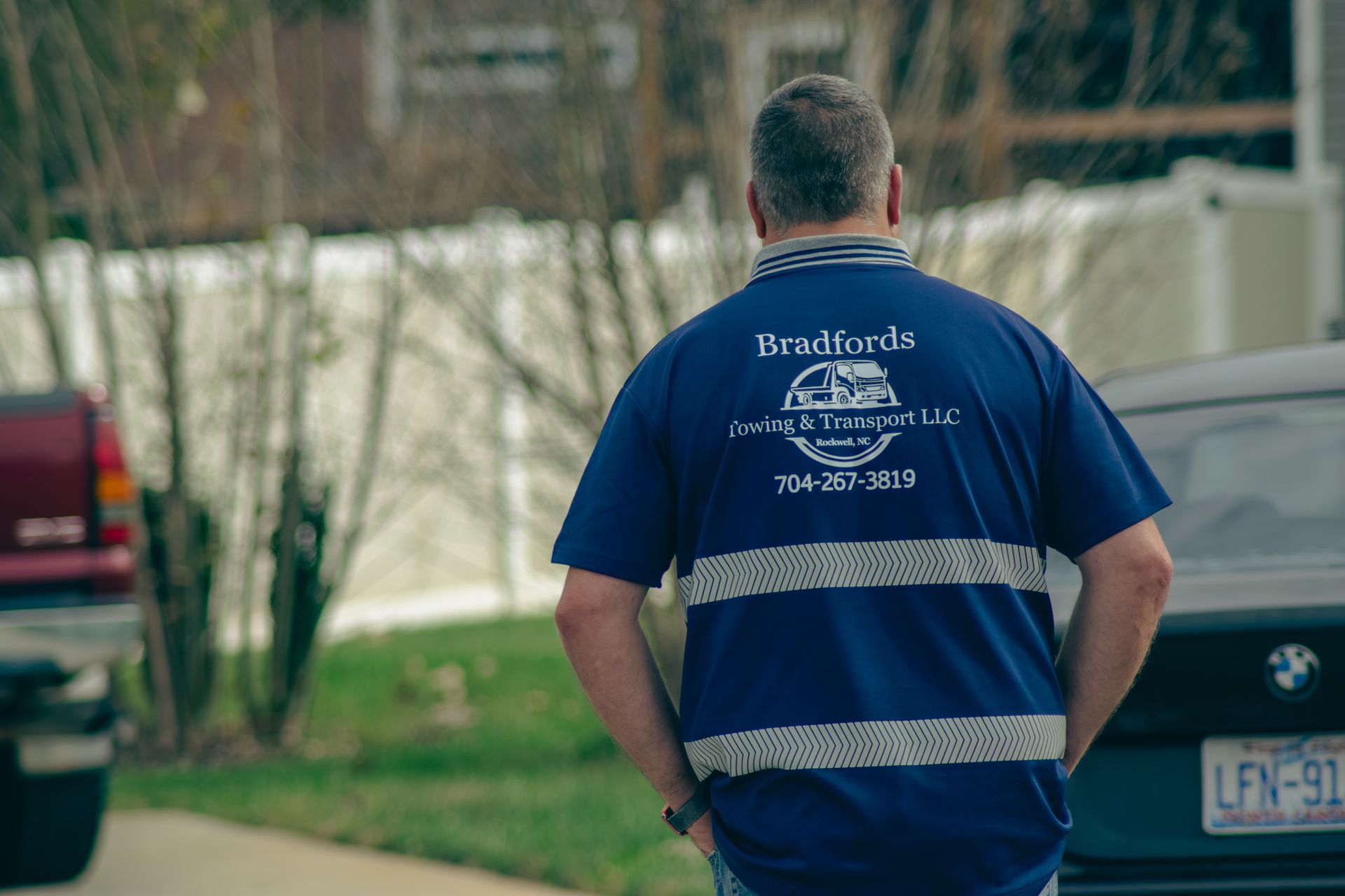 Person in a blue shirt with reflective stripes walking outdoors on a street near parked cars.