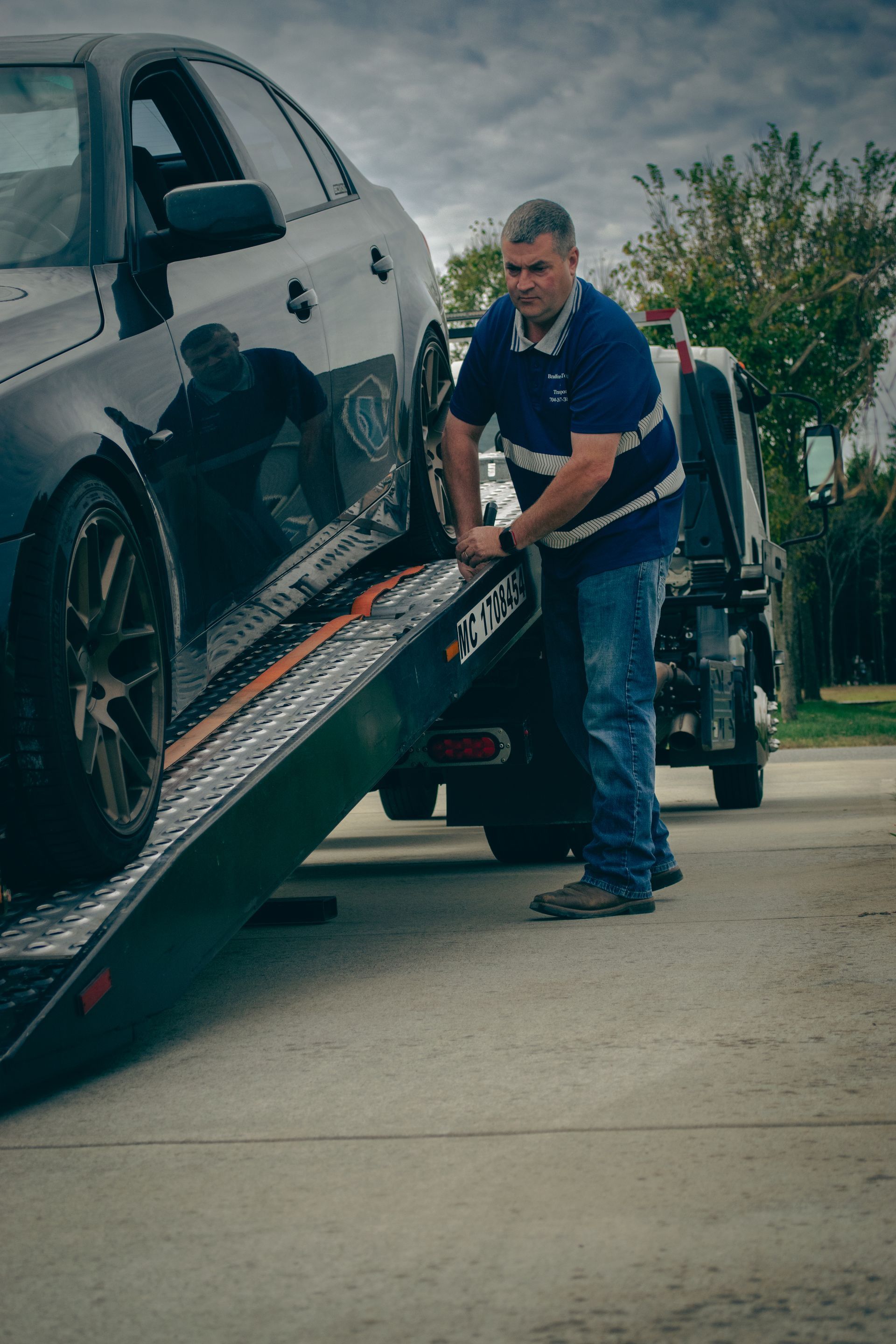 Tow truck operator securing a black car on a flatbed ramp outdoors