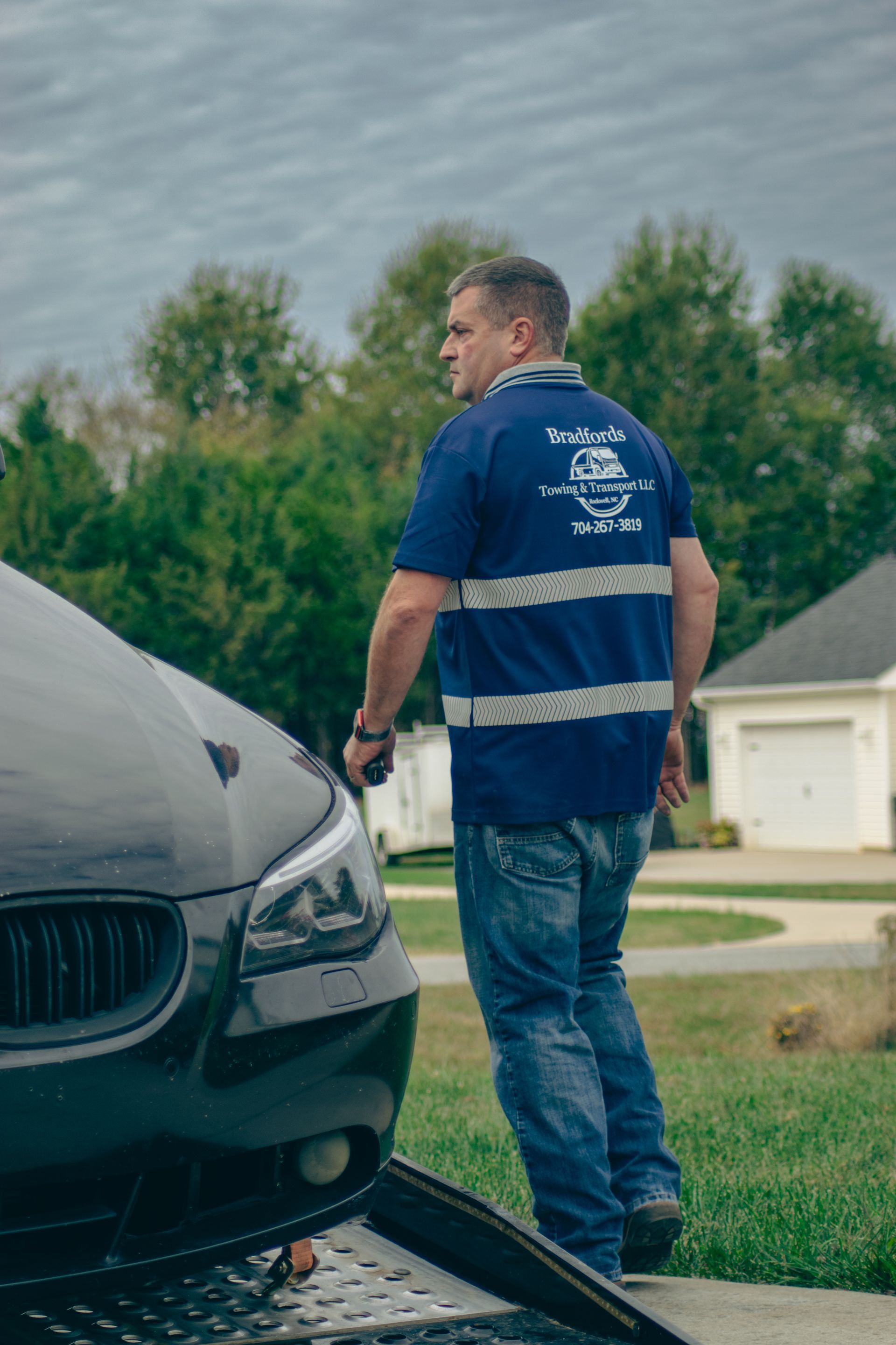 Man in blue shirt standing beside a black car on a grassy roadside near houses