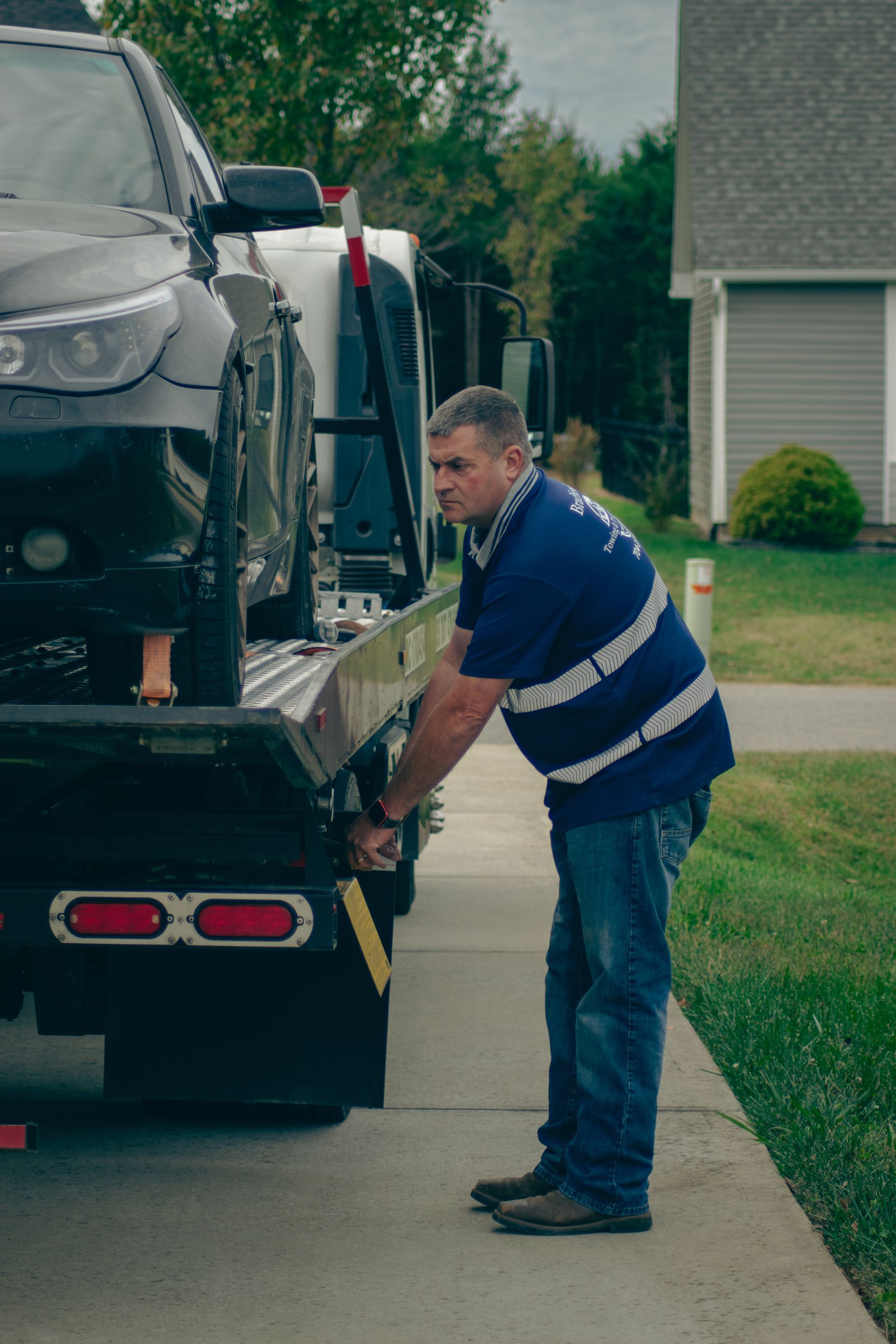 Tow truck driver securing a silver car on a flatbed in a residential driveway