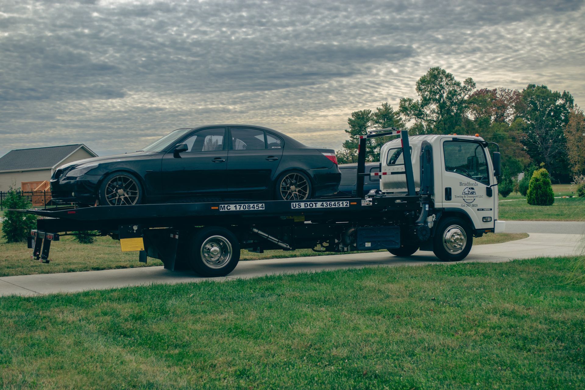 Black sedan loaded on a white tow truck in a grassy field under a cloudy sky