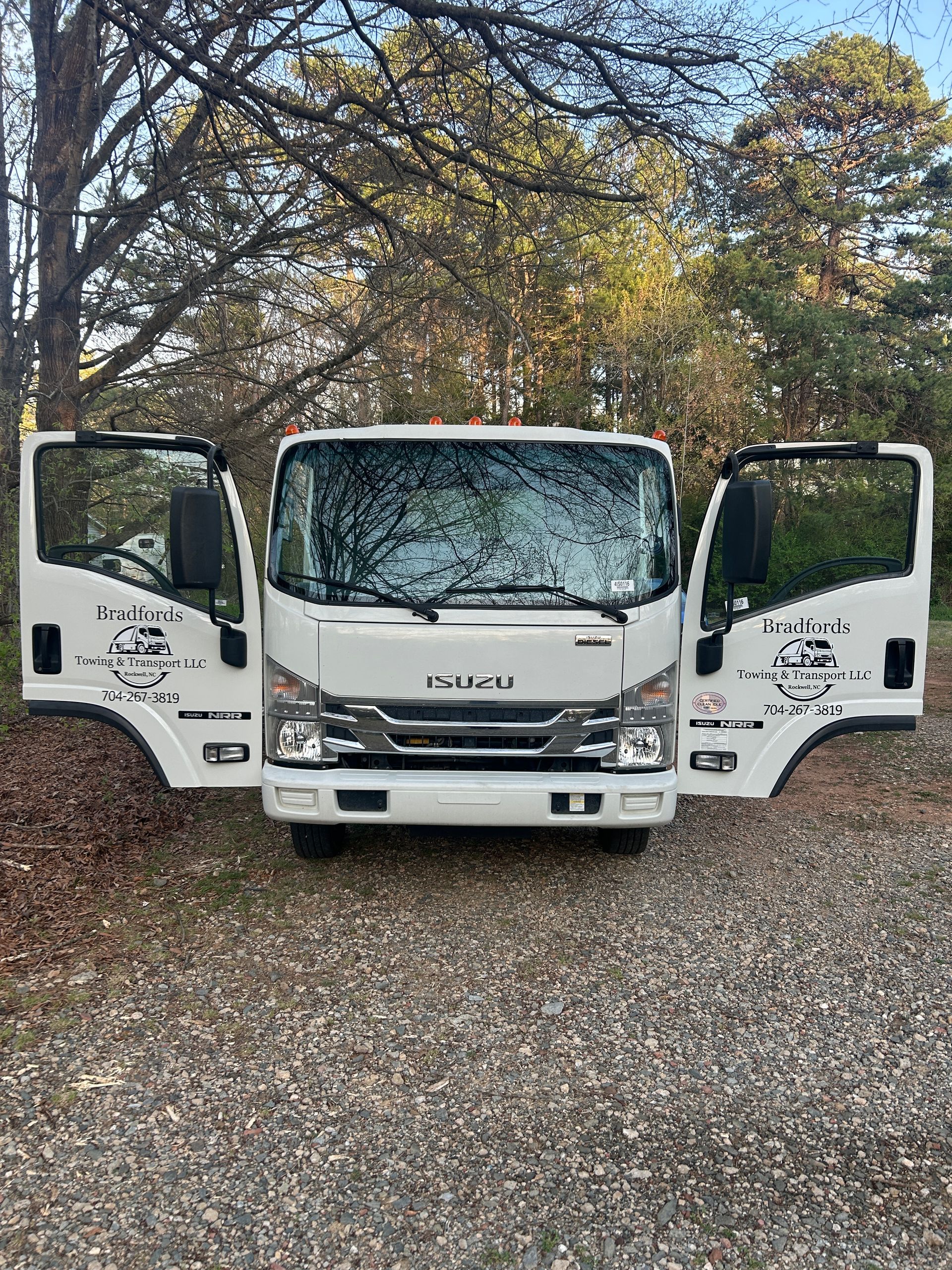 White Isuzu truck with both front doors open, parked on gravel beneath trees.