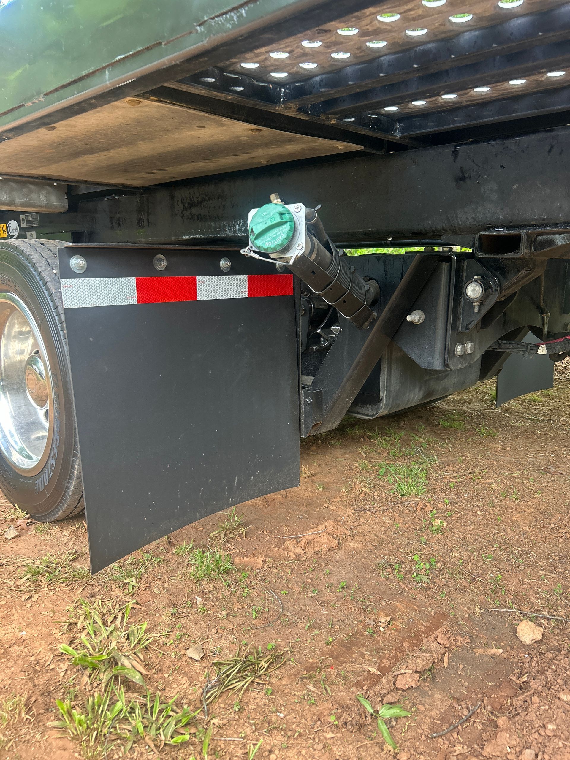 Underbody view of a trailer with a black mudflap and reflective red-white striping on gravel and grass