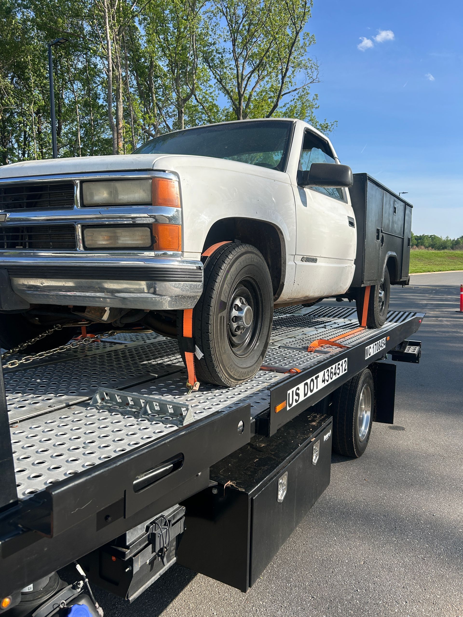 White pickup truck on a flatbed trailer, secured with orange straps, parked outdoors under a blue sky