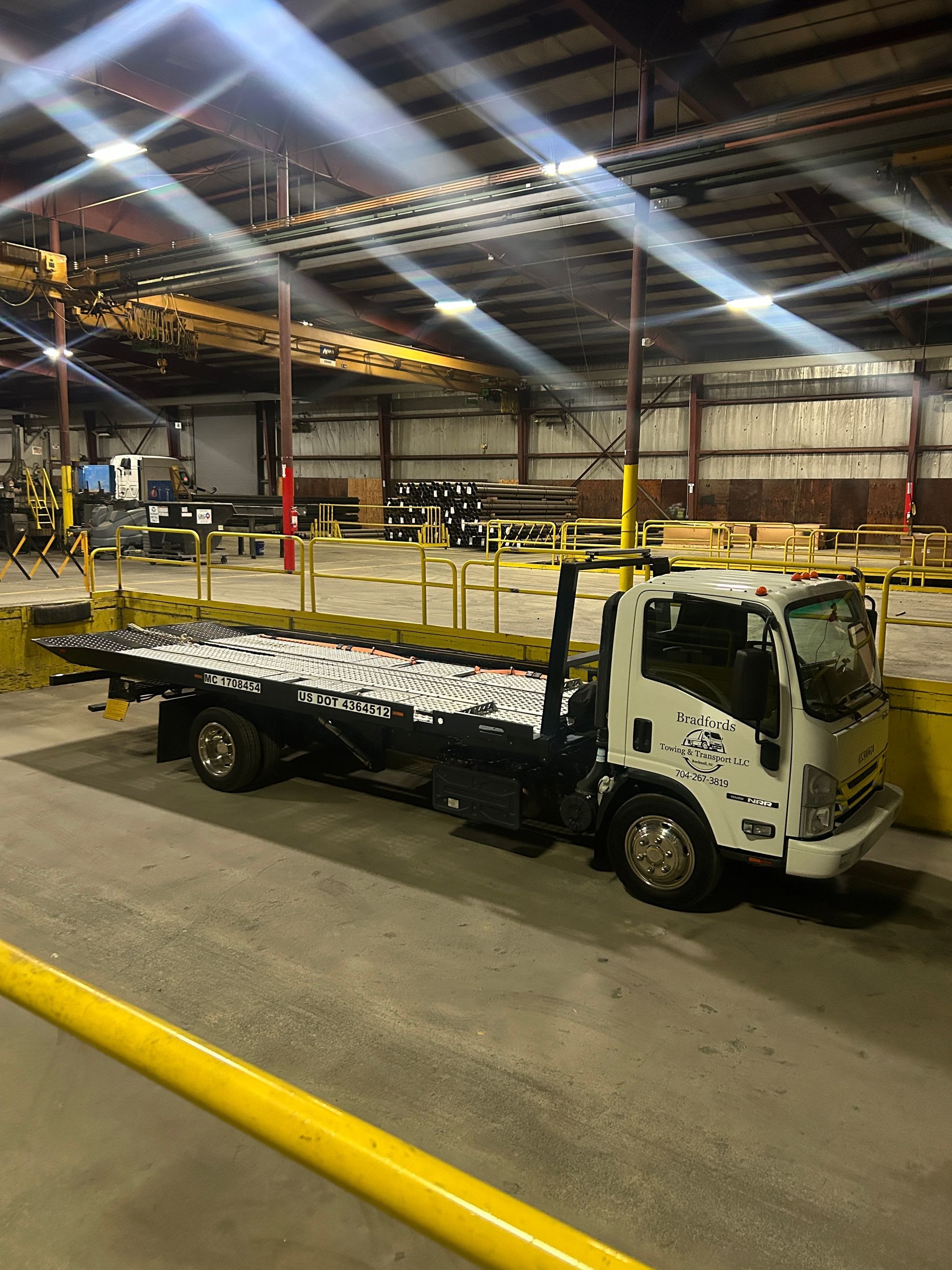 White flatbed truck loaded with wrapped pallets inside a warehouse, under bright overhead lights.