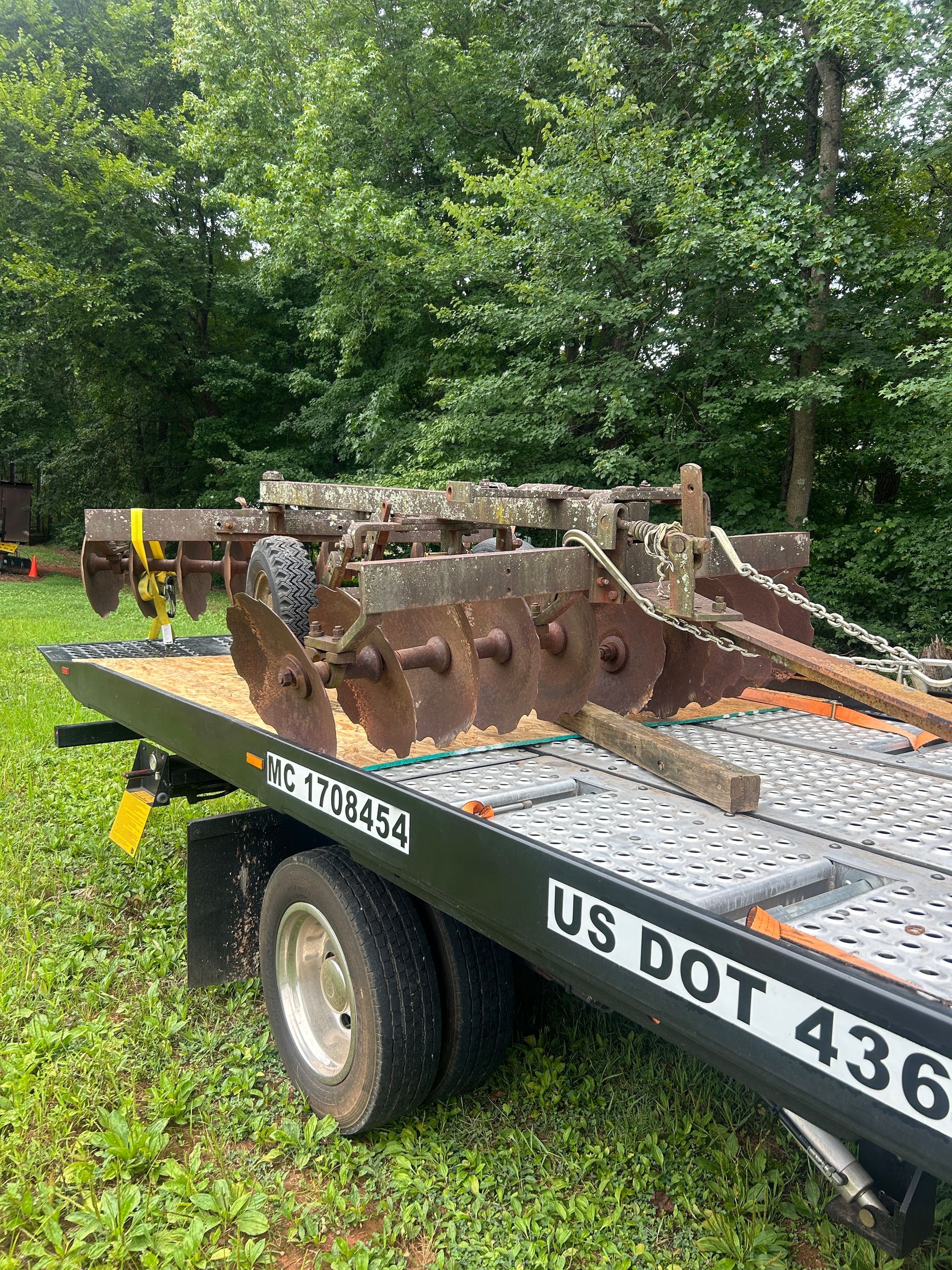 Rusty metal farm implement on a flatbed trailer in a grassy, wooded area.