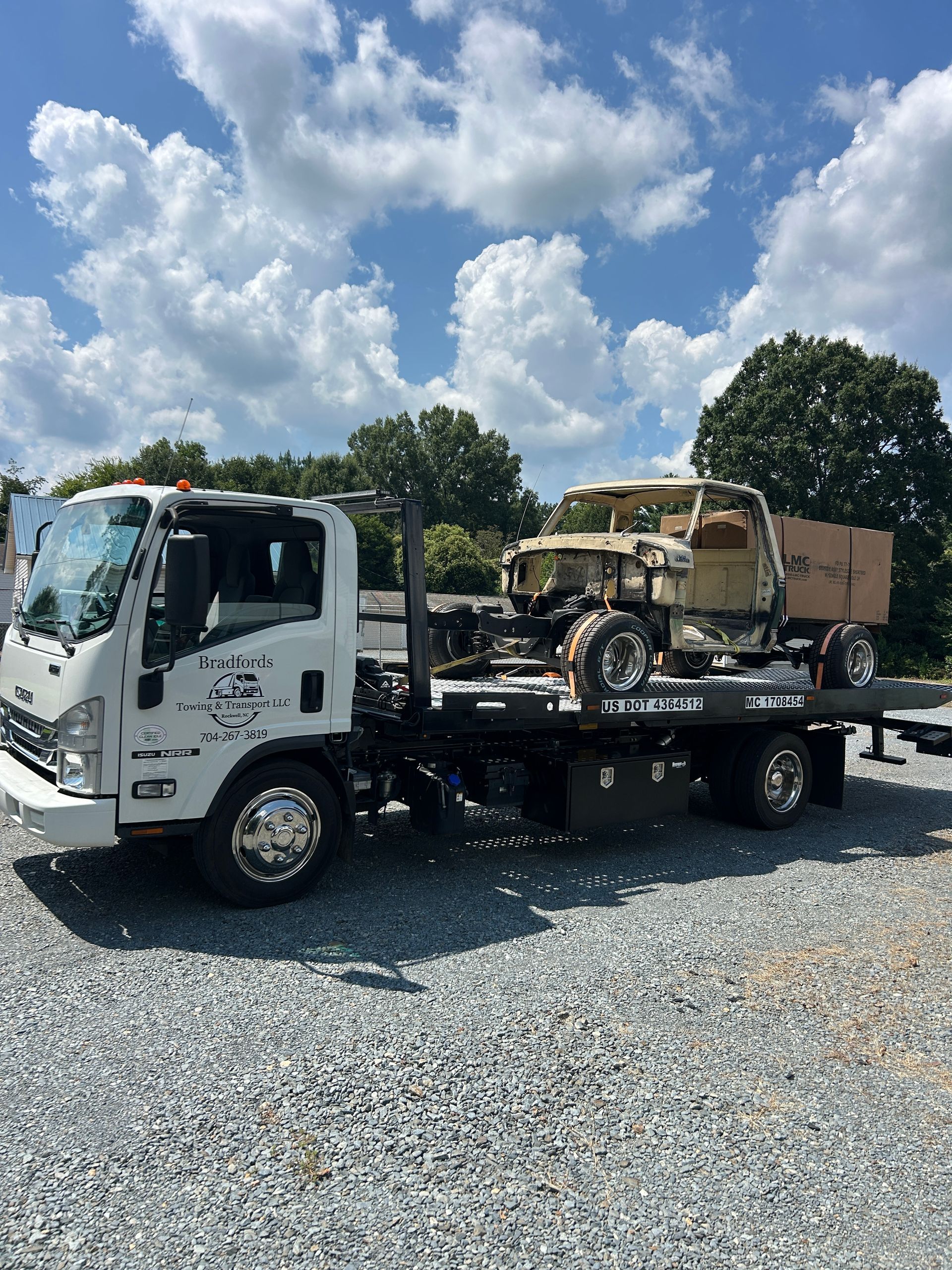 White tow truck carrying a tan off-road vehicle on a flatbed, parked on gravel under a partly cloudy sky