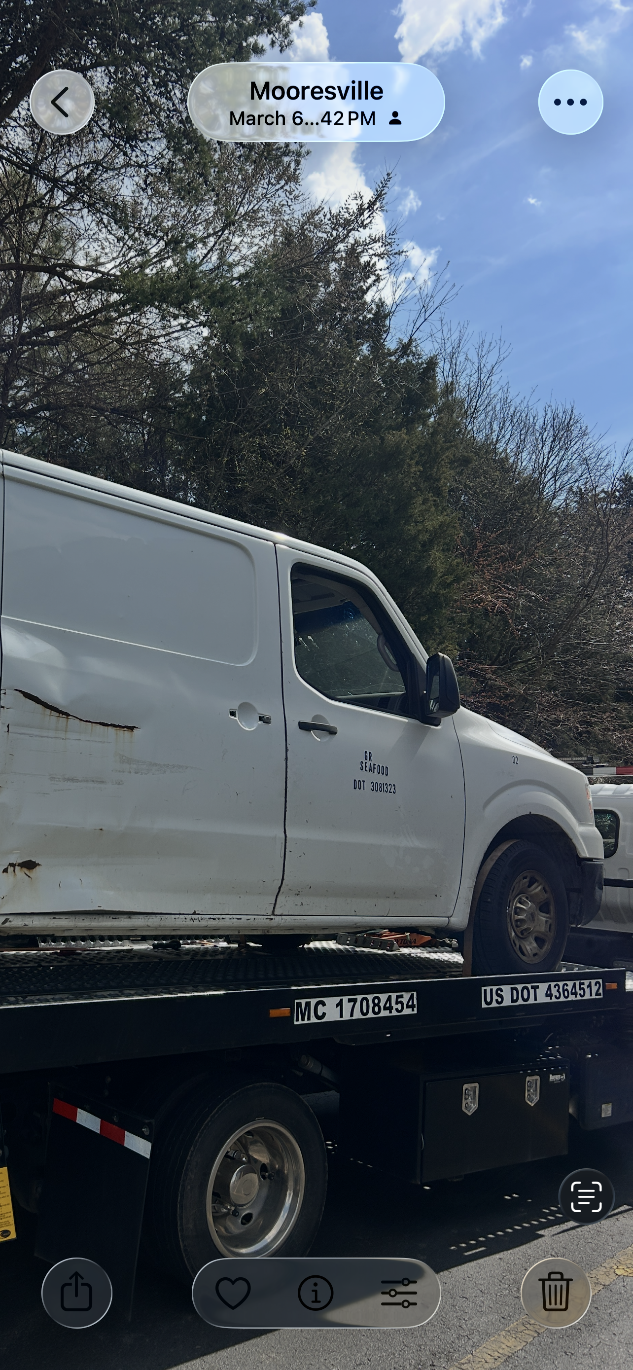 White truck on a flatbed tow trailer under a blue sky, viewed from the side on a tree-lined road