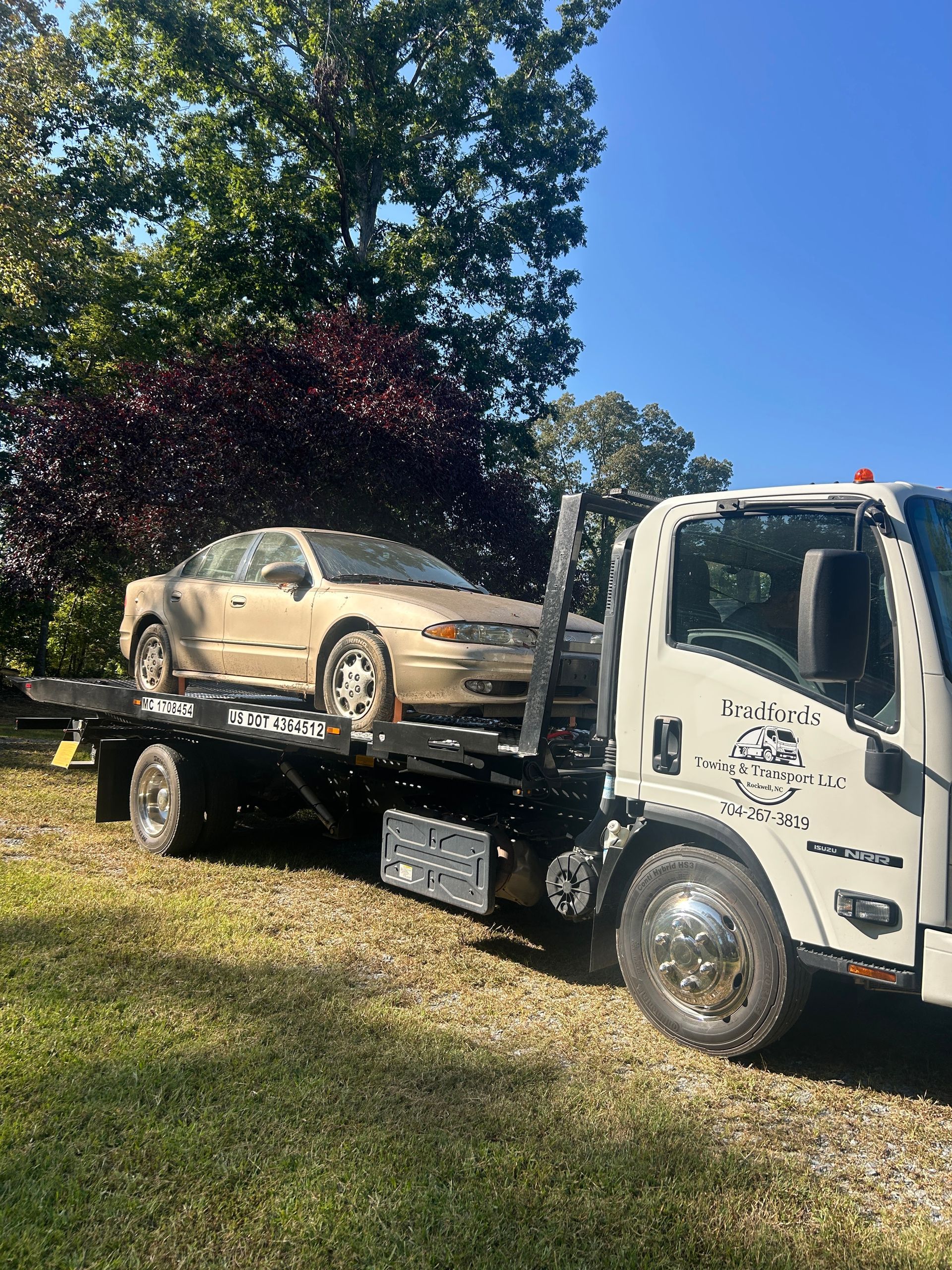 Tow truck hauling a beige car on a flatbed beside a grassy lawn and trees under a blue sky