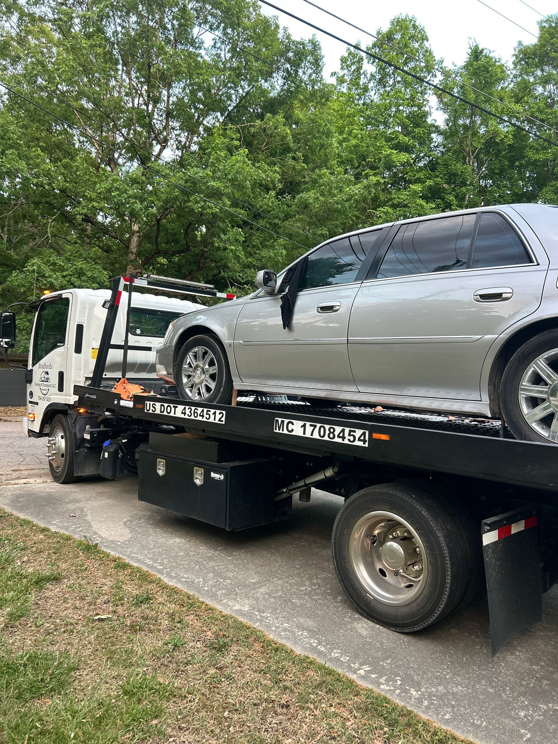 Silver sedan on a tow truck on a tree-lined street, with another truck behind it.