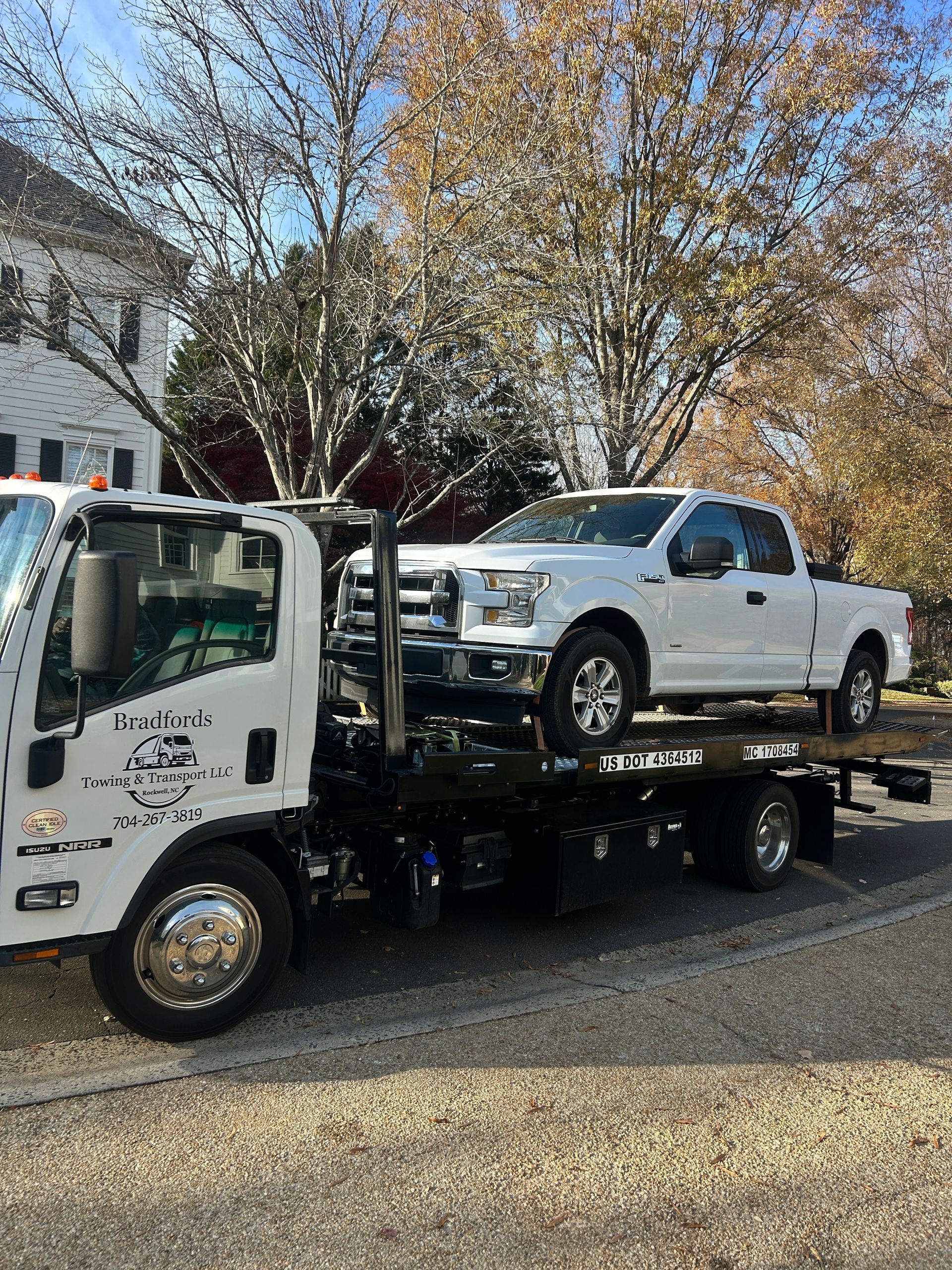 Tow truck carrying a white pickup in a driveway with trees and houses nearby