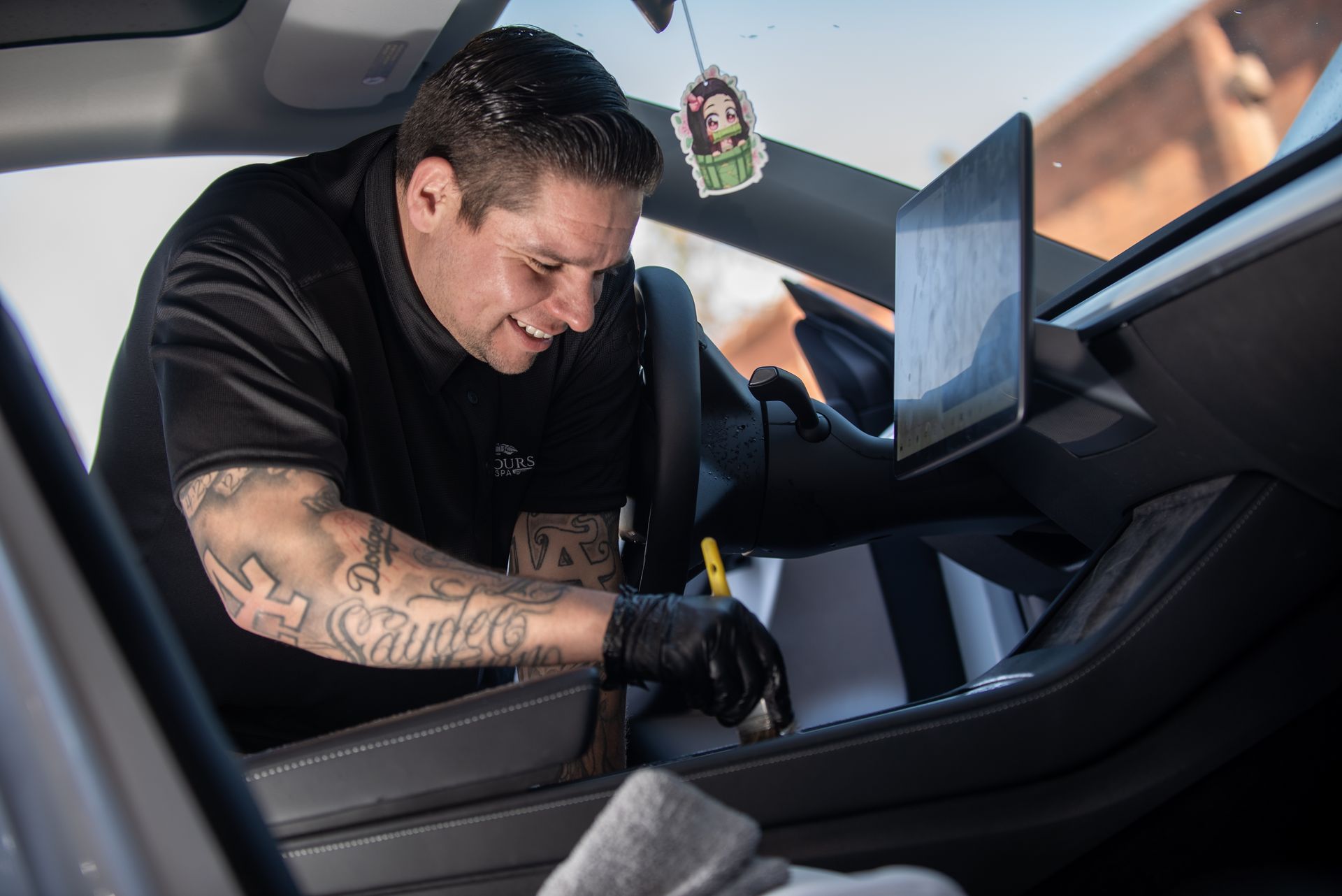 Man with tattoos cleaning a car's interior. He smiles, wearing black gloves, cleaning a dashboard with a brush.