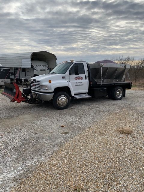 White dump truck with front plow parked on gravel near a barn under a cloudy sky