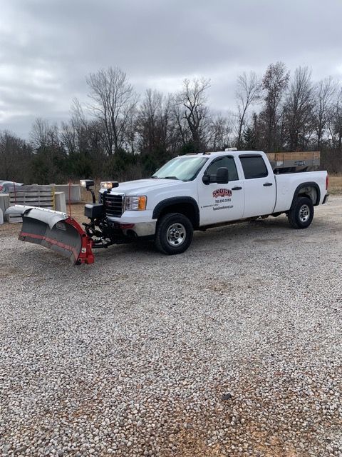 White utility truck with snowplow parked on gravel lot under cloudy sky.