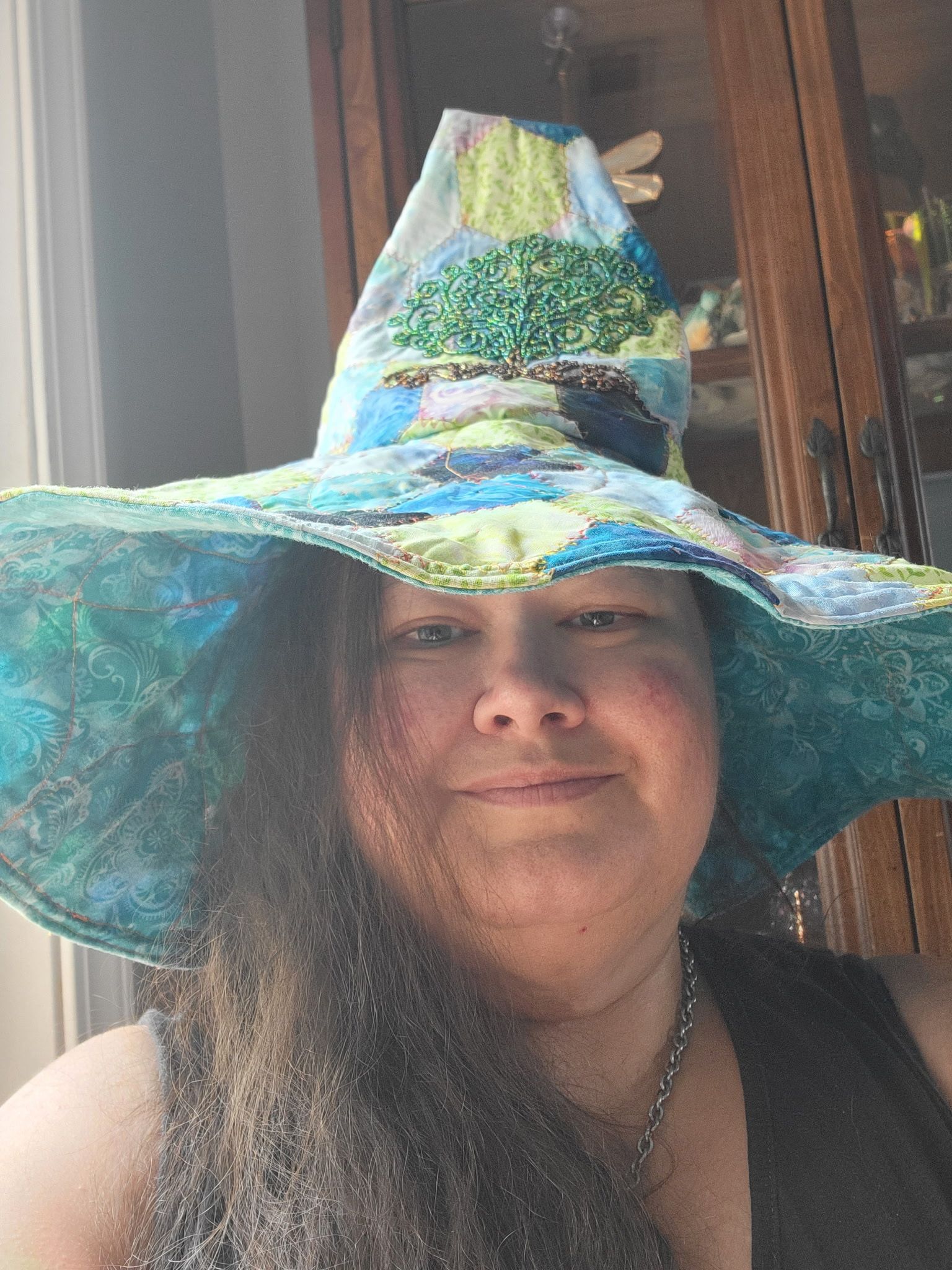 Person wearing a large blue-green wizard hat, smiling indoors near a wooden door.
