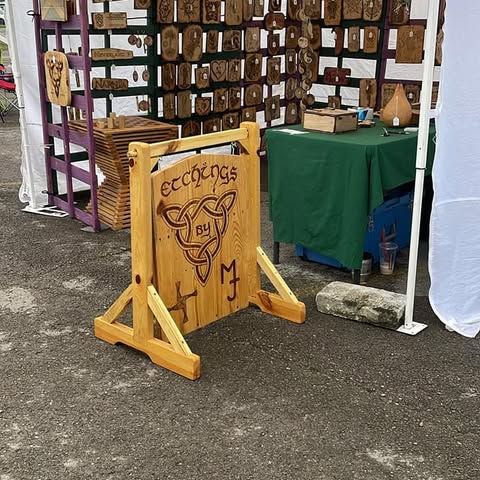 Wooden craft booth sign with carved ram logo beside a green-covered table at an outdoor market