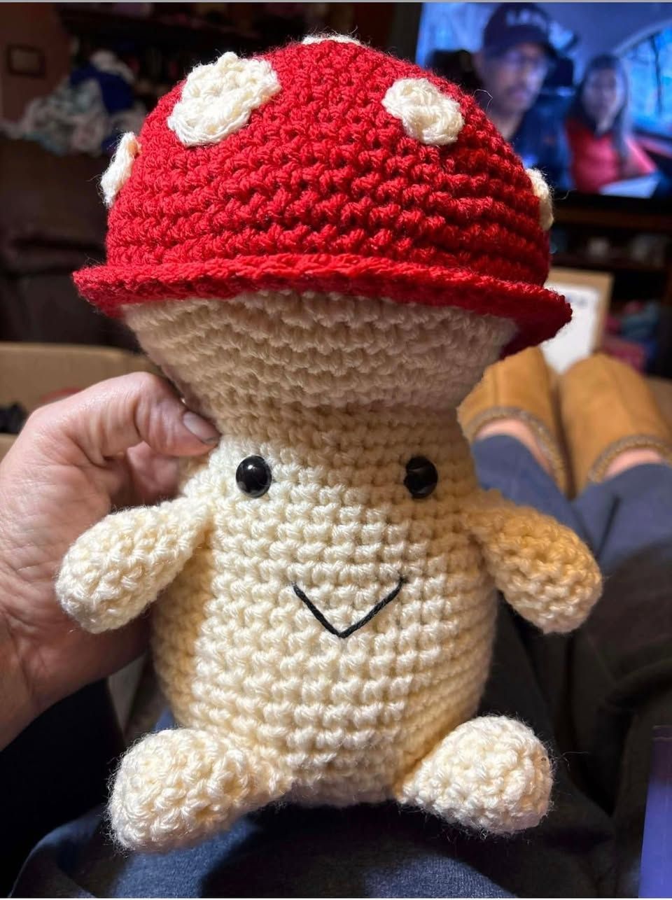 Crocheted mushroom plush with red-and-white spotted cap, beige body, and a small smiling face held in a hand