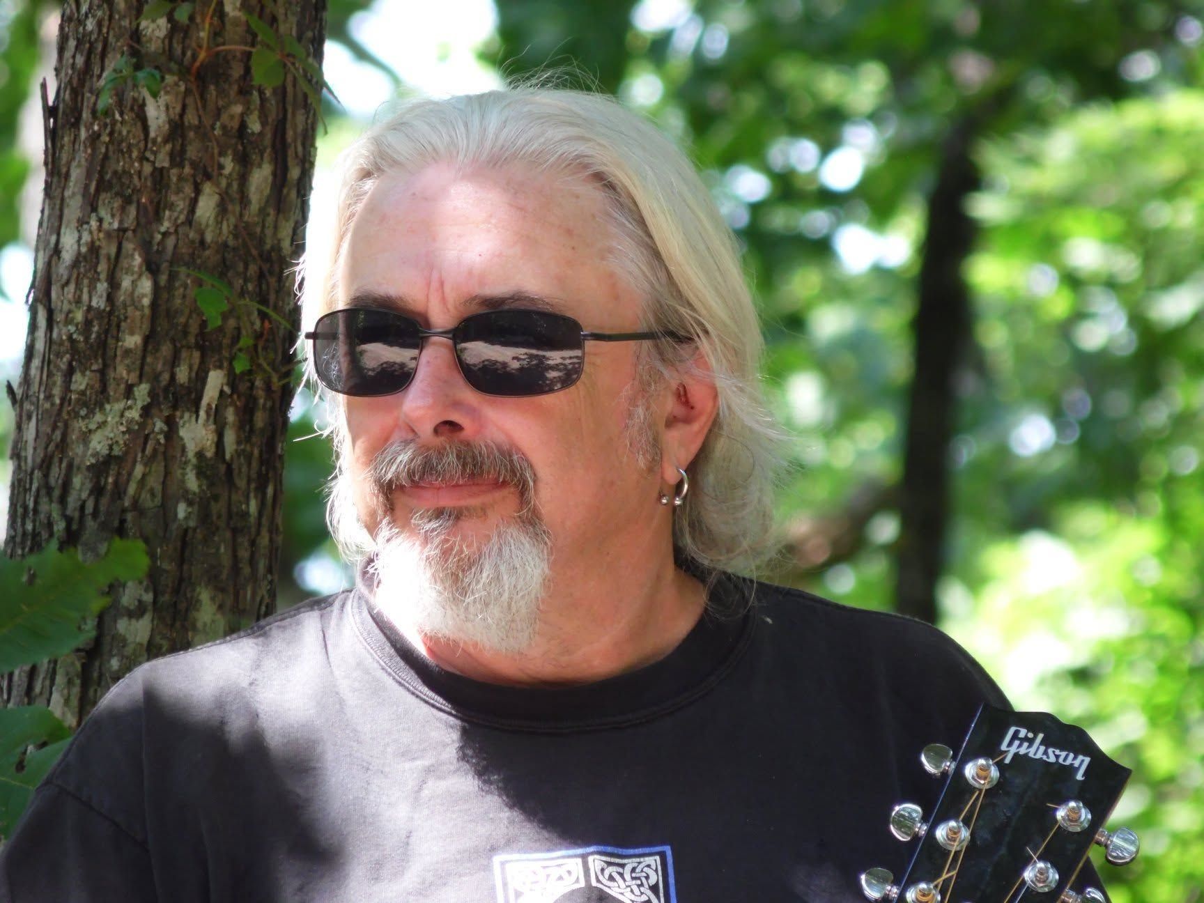 Man with sunglasses and gray hair standing by a tree, holding a guitar outdoors