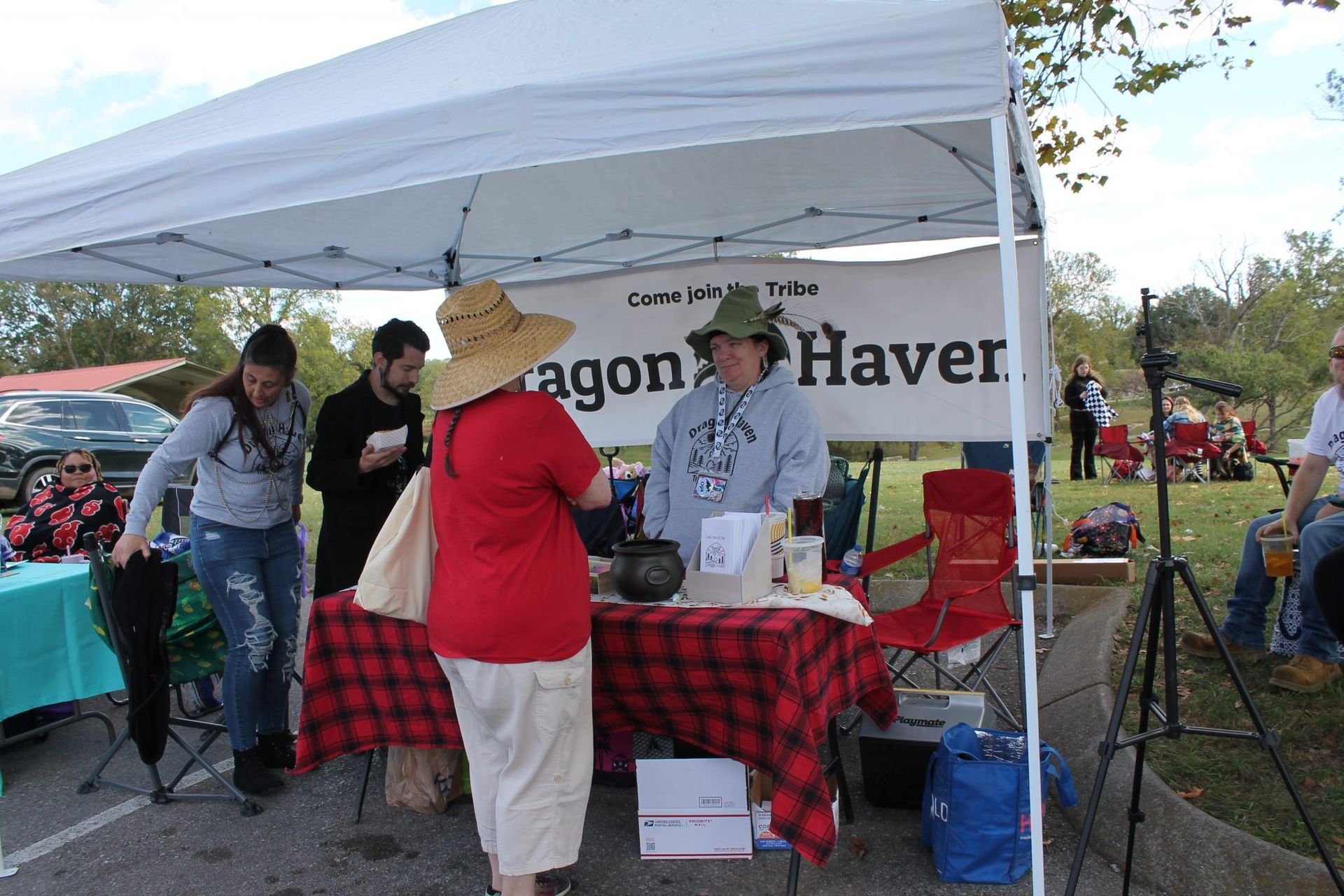 People at a Dragon Haven booth under a white canopy, with a red plaid tablecloth at an outdoor event