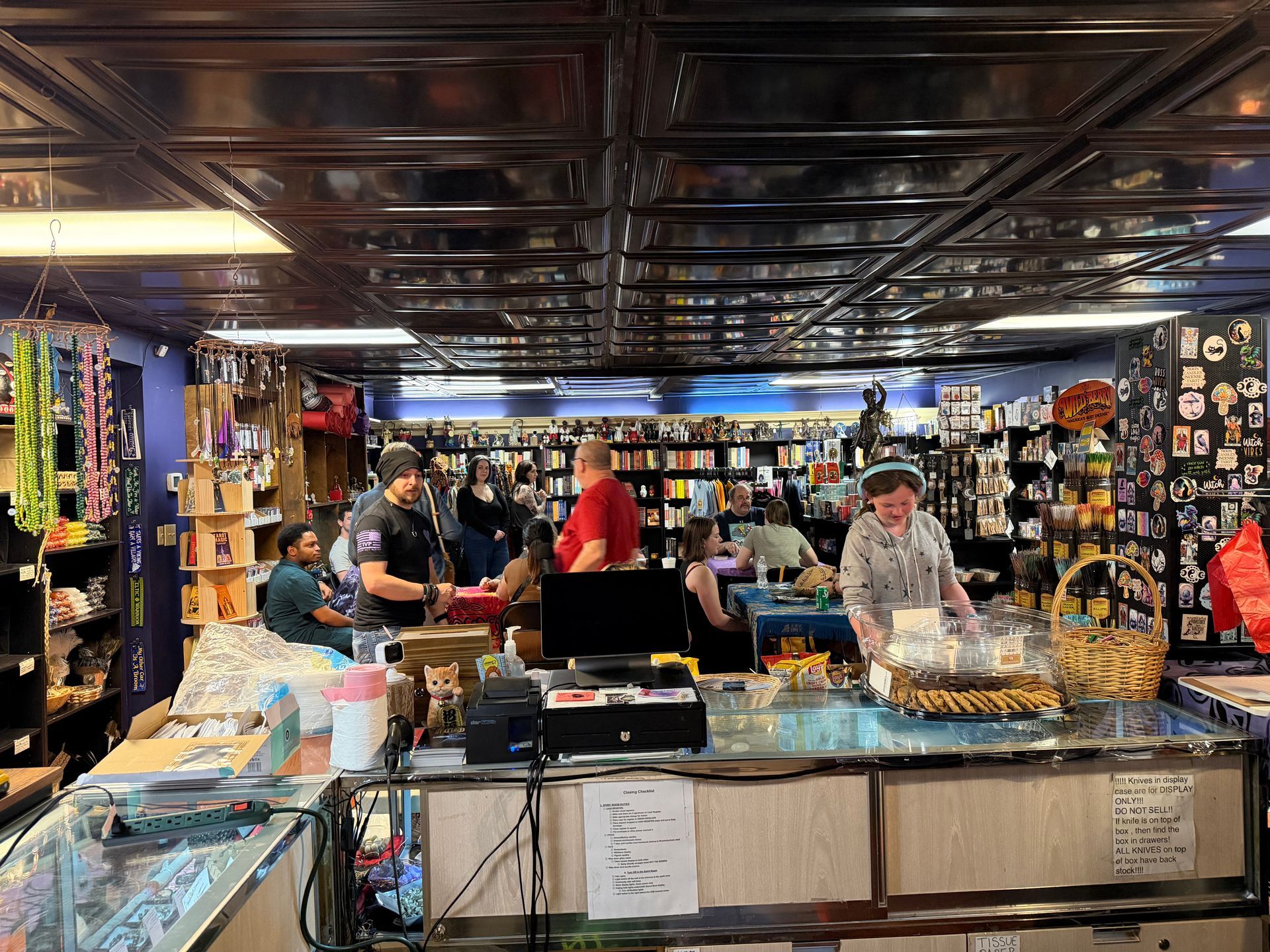 Busy indoor market stall with glass cases, hanging trinkets, and several people browsing under dark ceiling panels.