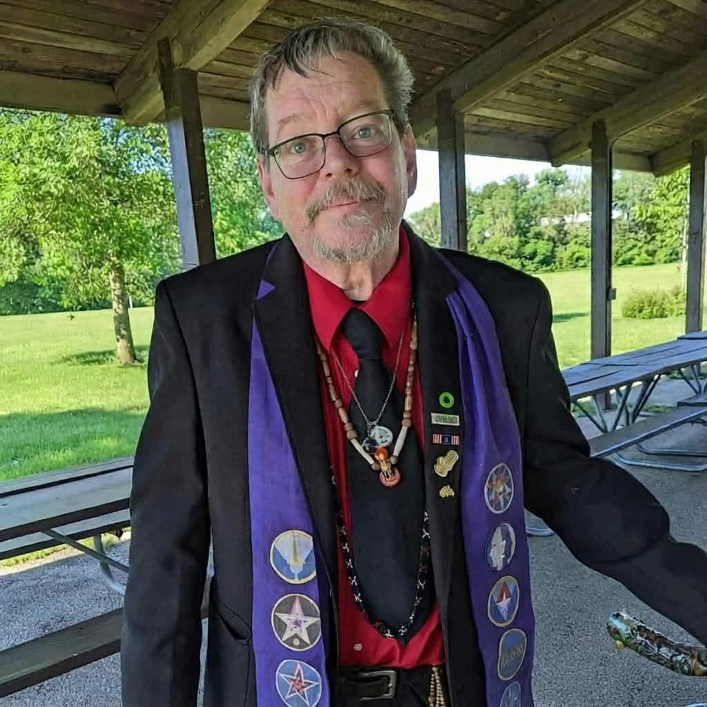Man under a pavilion wearing a black jacket with purple trim, red shirt, and medal ribbons, standing outdoors.