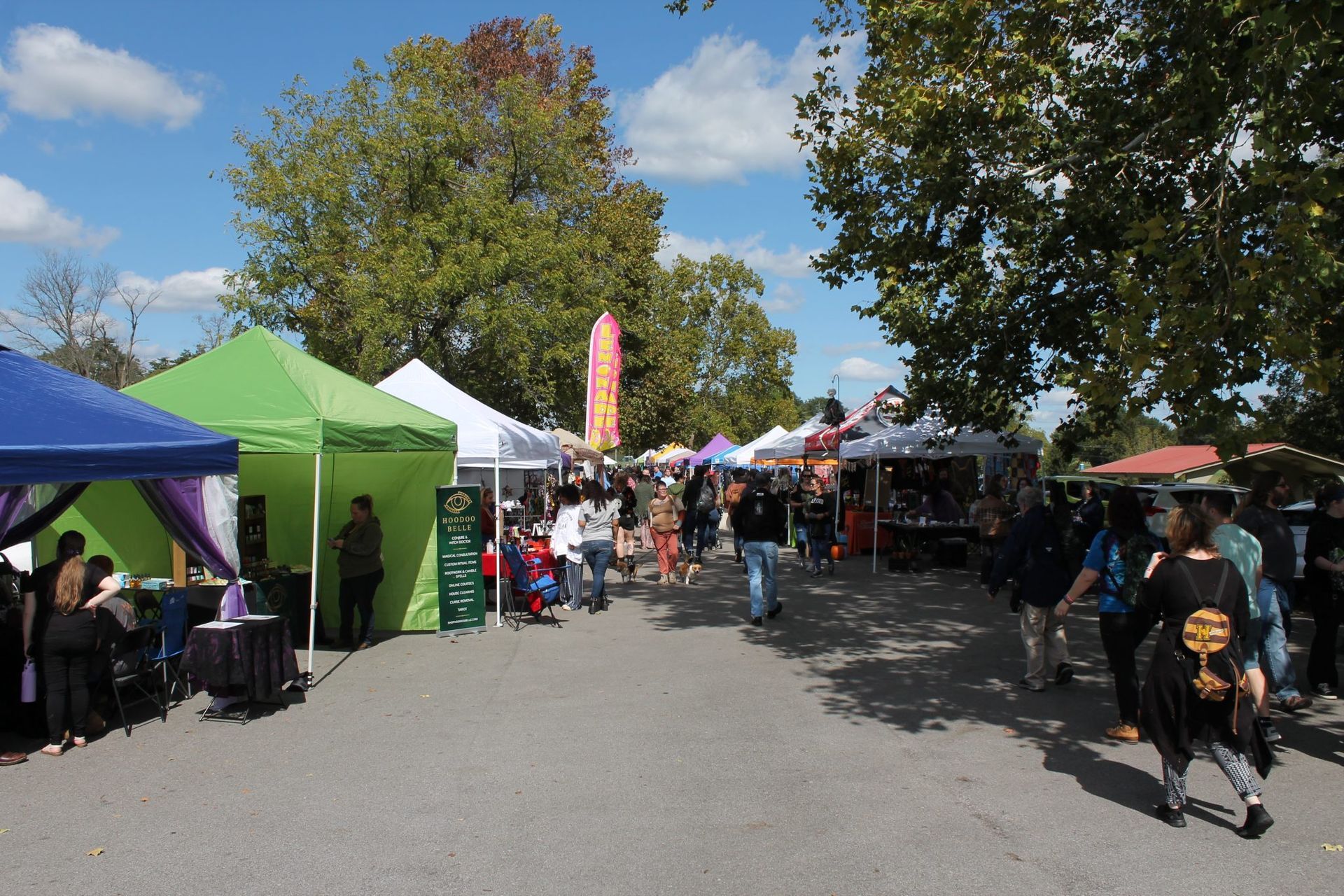 Outdoor market with colorful vendor tents, people browsing, and trees lining a sunny walkway.