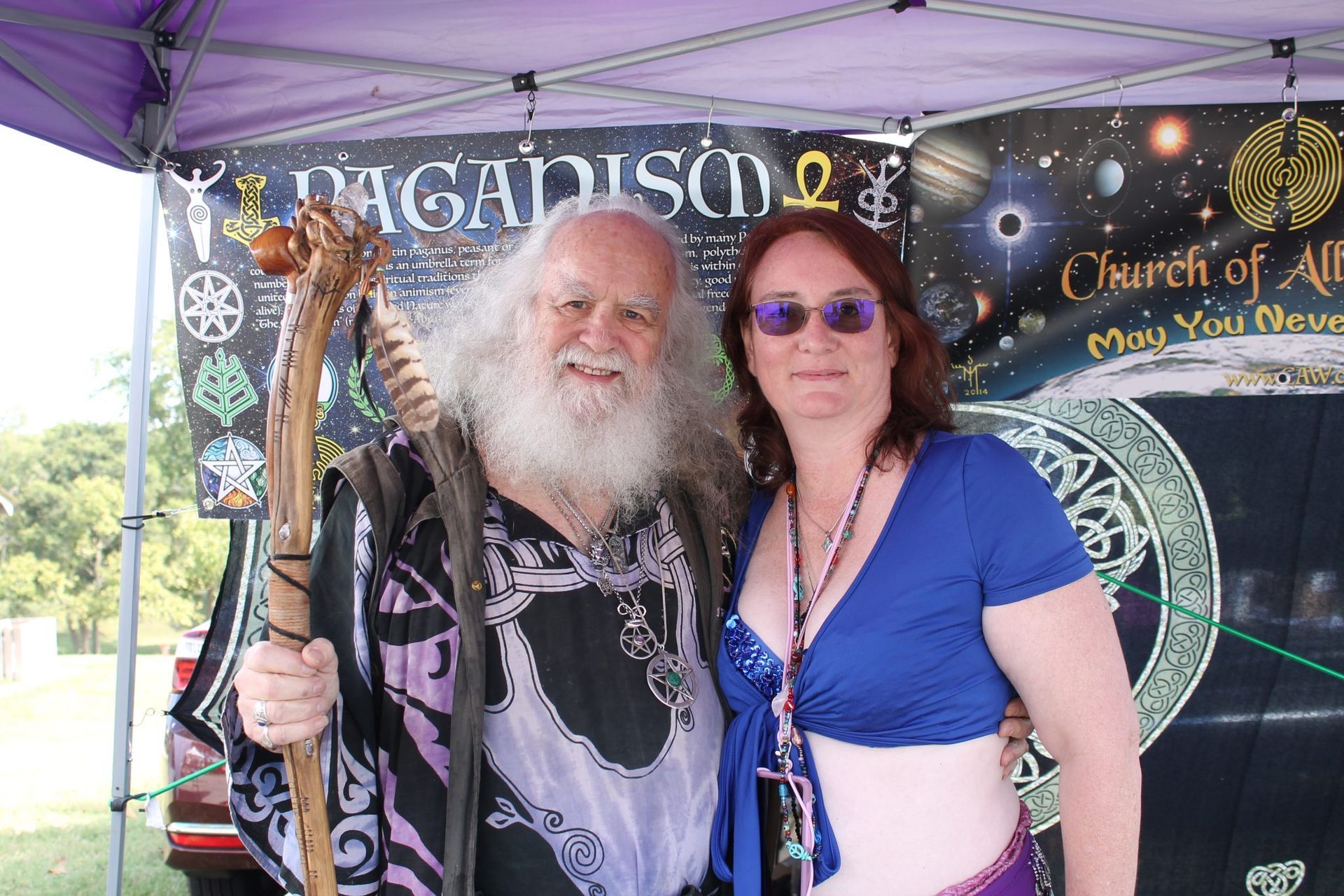 Two people posing under a purple tent at a pagan booth, one holding a staff, with a black banner behind them.