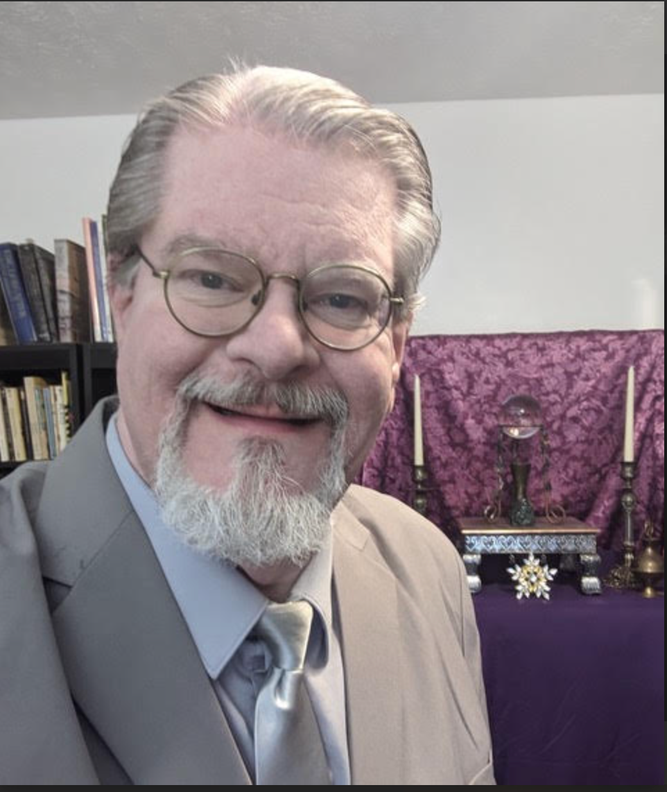Smiling man in glasses and gray suit in front of purple altar with candles and books