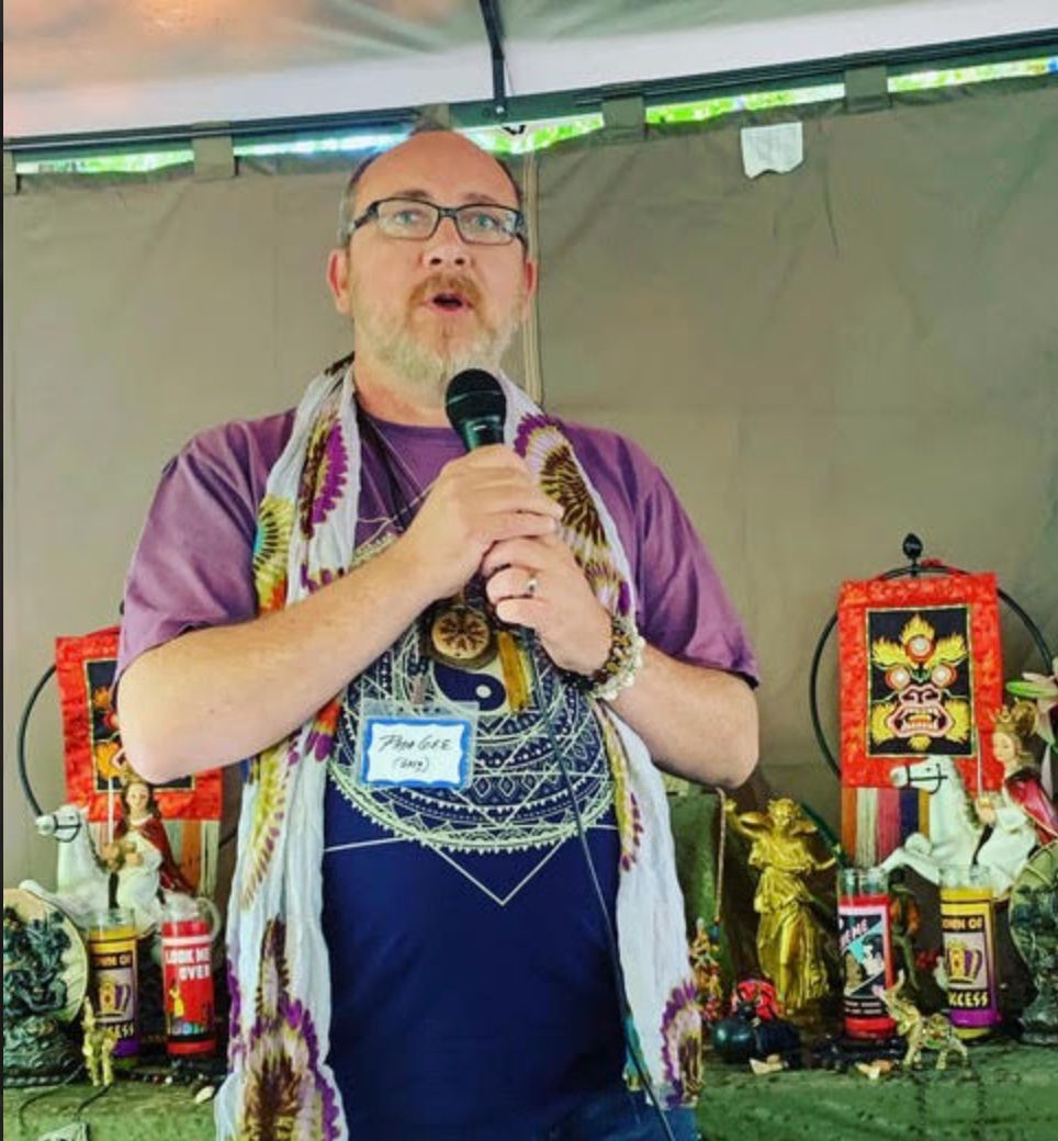 Man speaking into a microphone at a table with candles, trophies, and decorative items under a tent