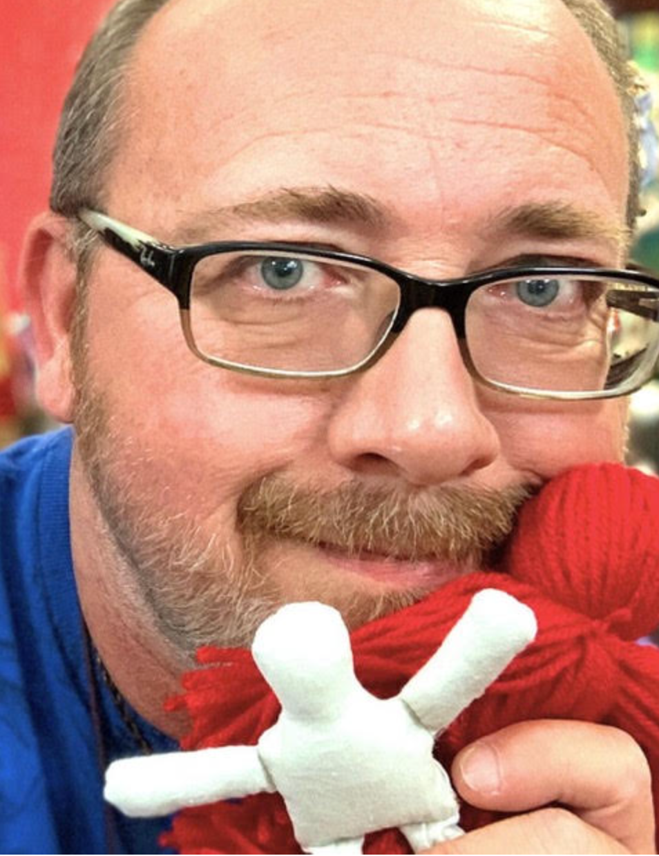 Man with glasses holding a small white puppet against a red background