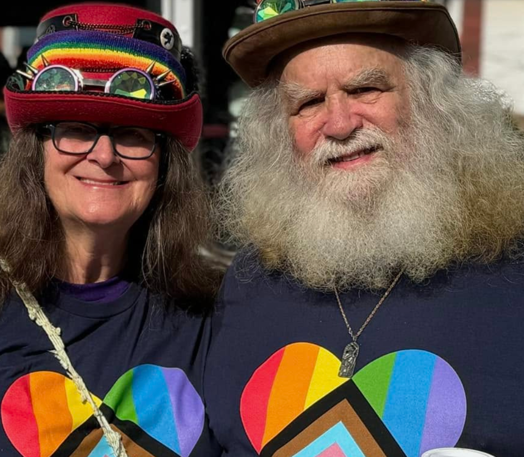 Two people in colorful rainbow shirts and hats smiling outdoors